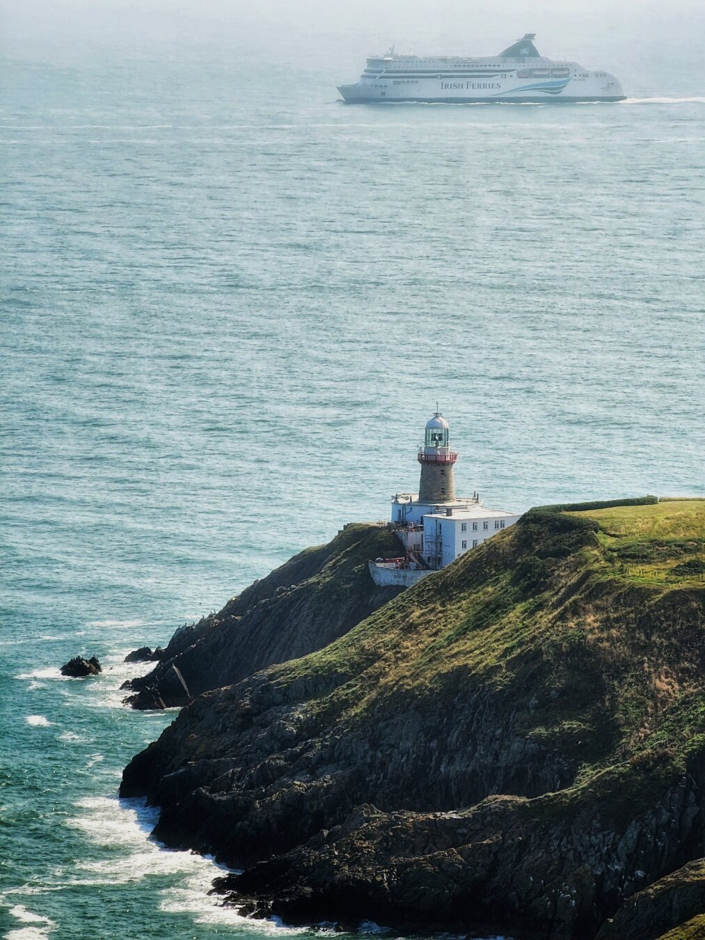 A vertical shot of an Irish lighthouse on a grassy cliff, with a large white ferry from Irish Ferries sailing across the sea in the background. The sky is hazy and the water is a deep blue-green, with white waves crashing on the rocks below.