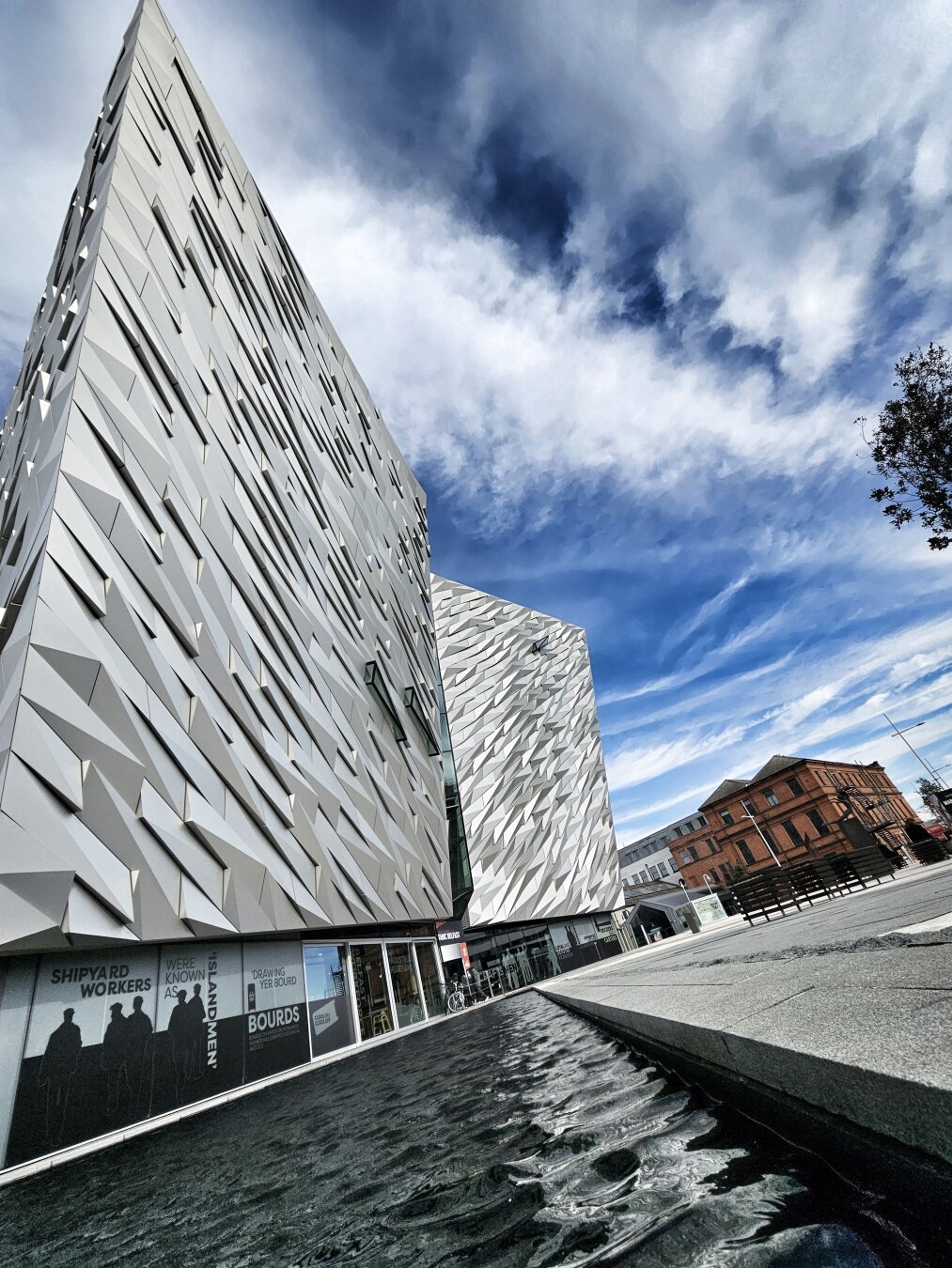 A low-angle, wide-shot of the Titanic Belfast building on a sunny day with a dramatic blue sky and wispy white clouds. The building's angular, silver-grey, textured facade dominates the left side of the frame, with its reflective surface catching the light. In the foreground, a dark pool of rippling water reflects the building. To the right, a street recedes into the distance, with a traditional red-brick building visible further down.