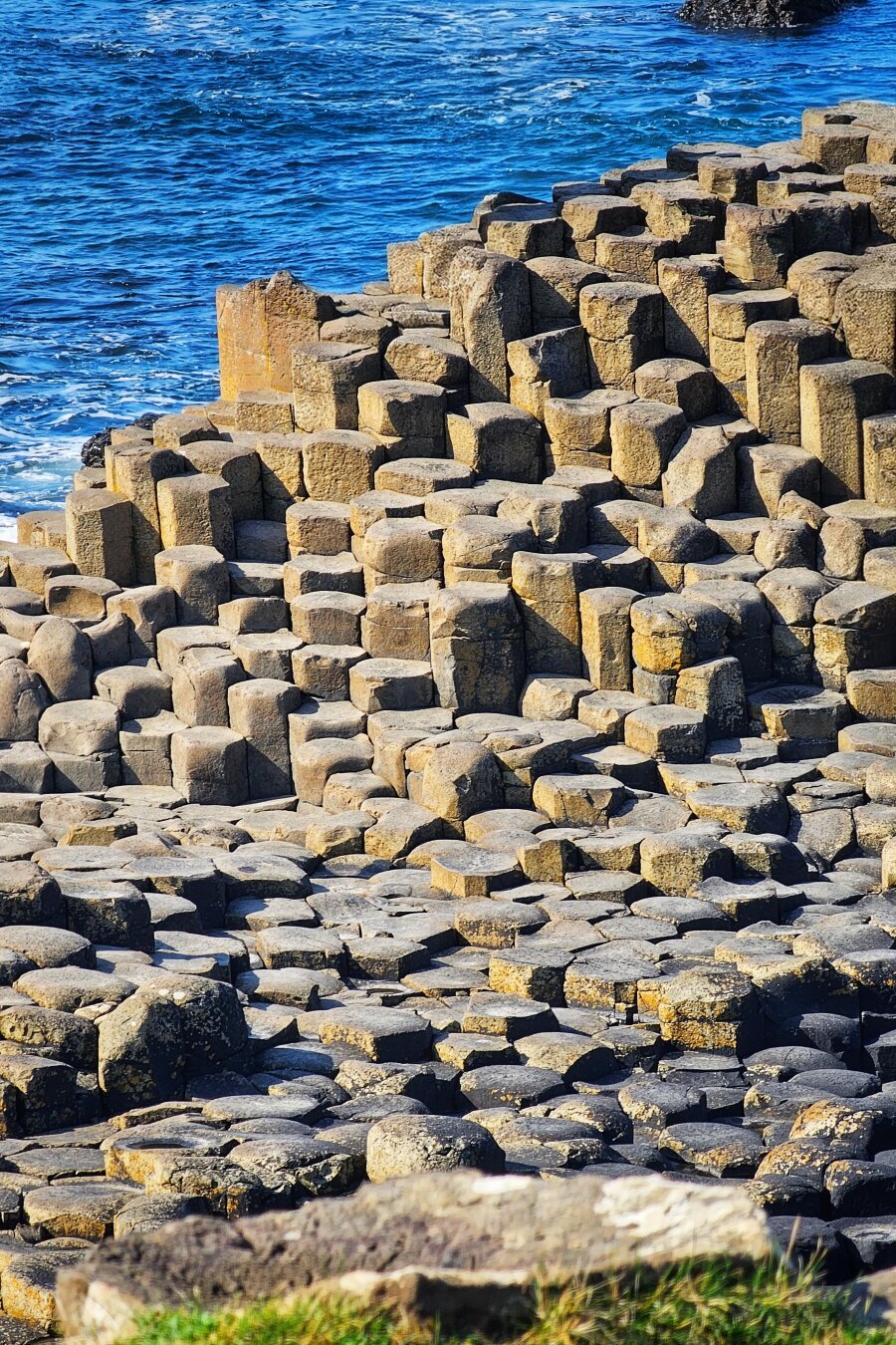 A close-up vertical shot of the hexagonal basalt columns of the Giant's Causeway in Northern Ireland. The dark grey and brown columns vary in height, creating a textured, stepped landscape that meets the vibrant blue, slightly frothing ocean at the top left of the frame. Some green foliage is visible in the blurred foreground.