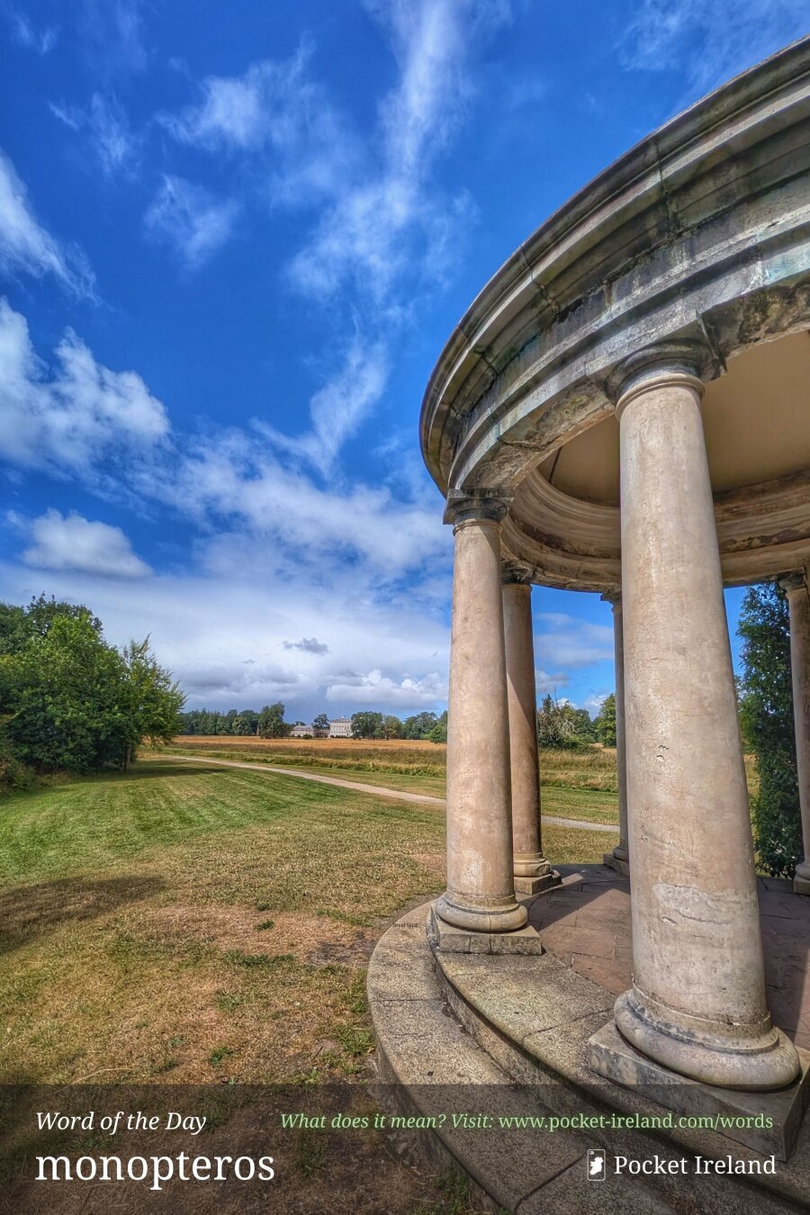 A low-angle view from beneath the weathered stone pillars of an open, circular classical rotunda. Pale cylindrical columns and a curved entablature frame the right foreground, casting soft shadows across a stone plinth. Beyond the pavilion, a dirt track curves across sun-drenched green and tawny parkland towards a stately country mansion in the far distance, set beneath a brilliant azure sky scattered with bright white clouds.