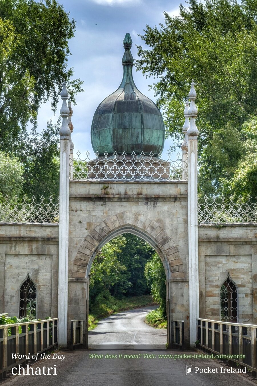 A stone gatehouse entrance featuring a central pointed archway, topped with an ornate, patinated copper onion dome and decorative white cresting. A narrow asphalt road passes over a bridge with metal railings directly through the arch into a sunlit, winding woodland path. Flanking stone walls with lancet windows are framed by lush, towering green deciduous trees beneath a blue sky with soft white clouds.