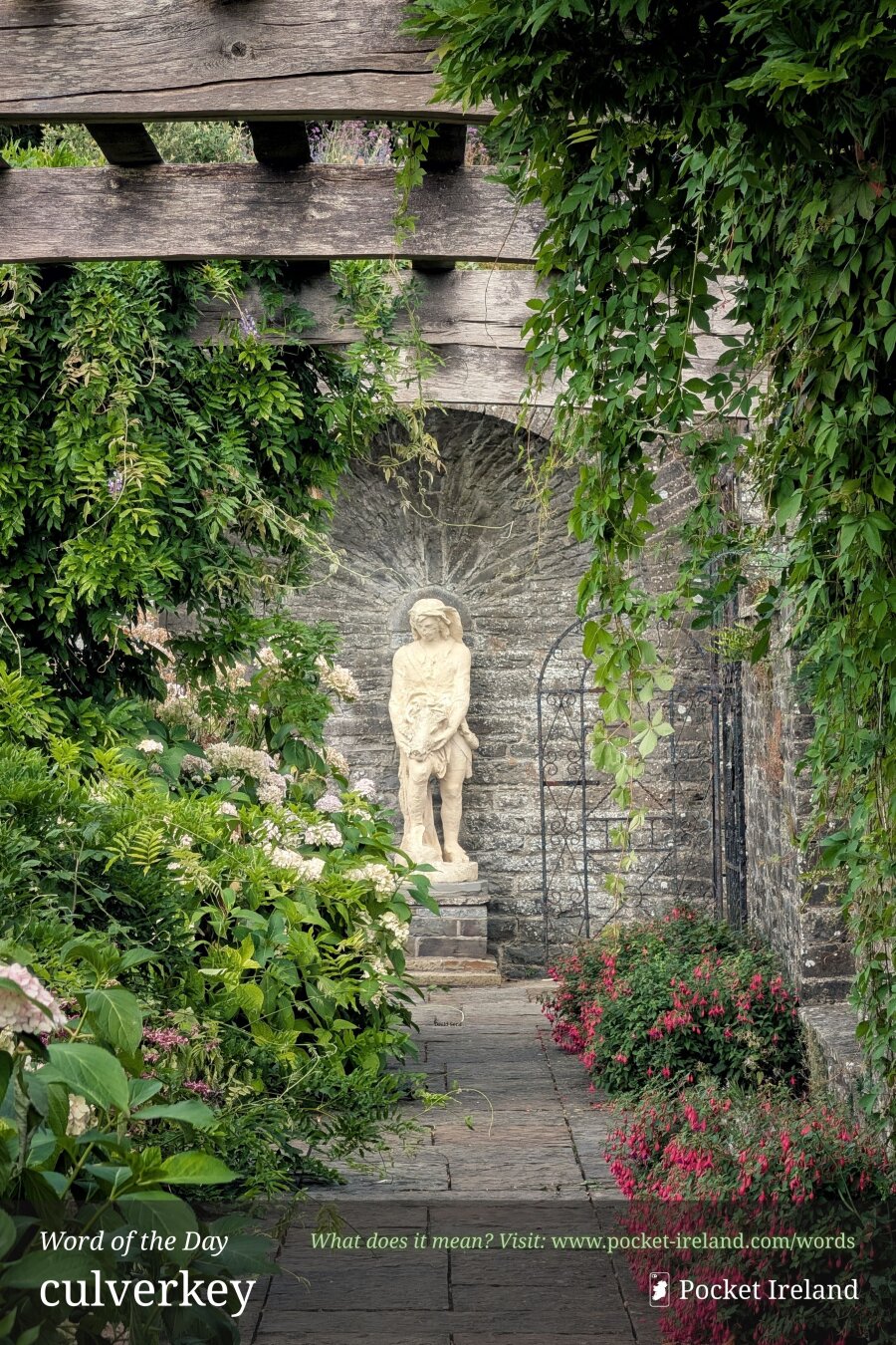 A stone-paved garden walkway framed by a weathered wooden pergola and lush climbing foliage. At the end of the path stands a classical pale stone statue of a figure set into an arched alcove of rough stonework. Flanking the path are flourishing hydrangea bushes with pale blossoms on the left and vibrant pink fuchsia flowers along the right. Trailing vines and leafy creepers hang gracefully overhead, creating a tranquil, sheltered garden sanctuary.