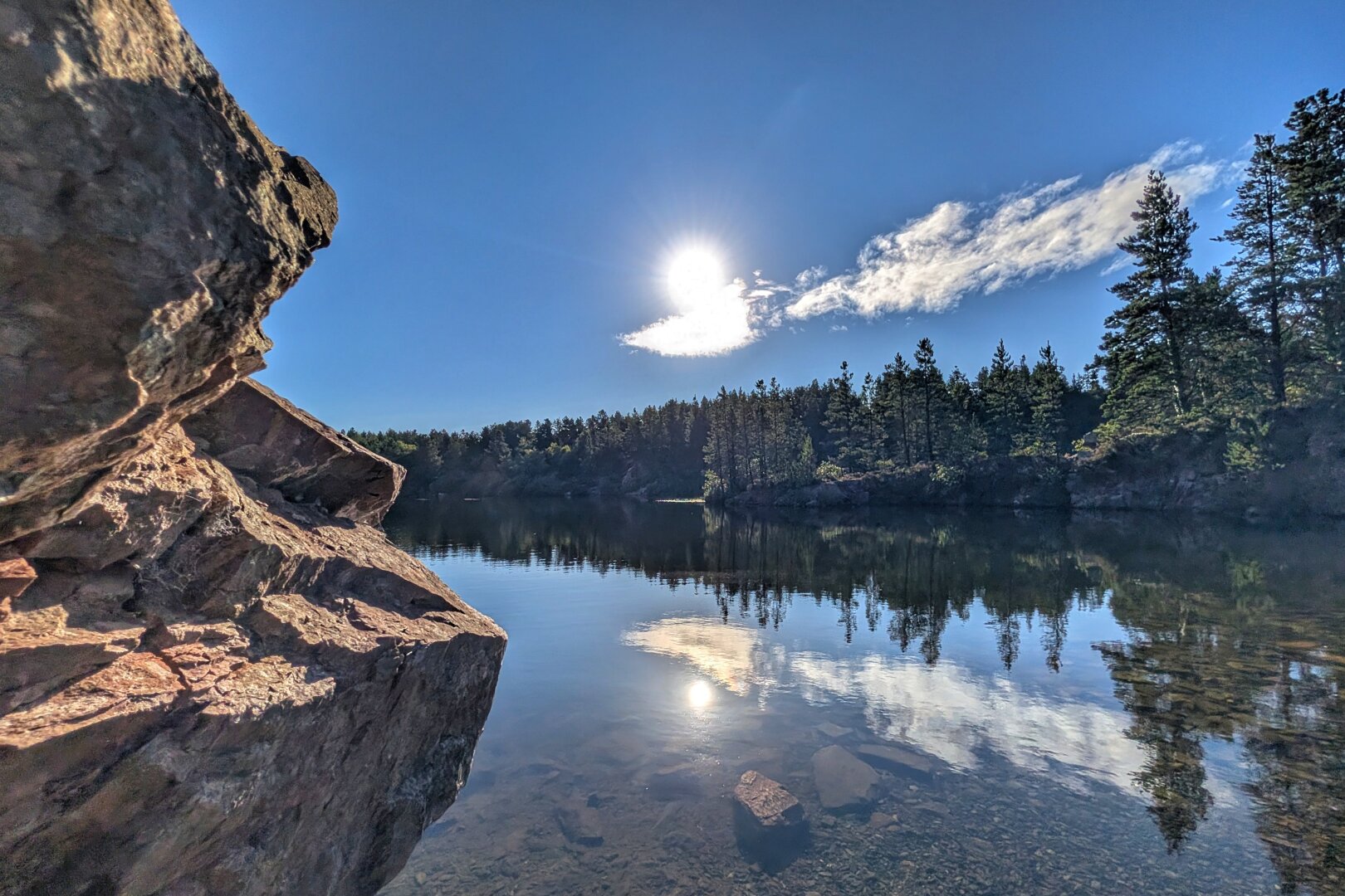 A wide shot of a tranquil lake on a bright morning. In the foreground on the left, a textured, warm-toned rock face is illuminated by sunlight. Crystal-clear, calm water reflects the vibrant blue sky, light clouds, and a dense line of pine trees that line the distant shore. The sun shines brightly above, casting a soft glow and mirror-like reflection across the gentle ripples of the water.