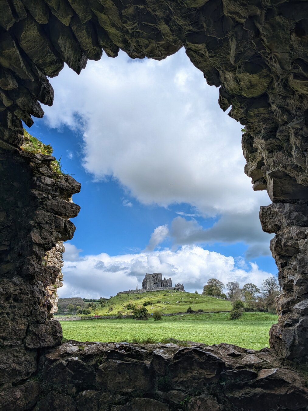 A vertical shot taken from inside a dark, rugged stone ruin, looking out through a jagged natural opening in the wall. In the centre distance, an ancient stone fortress sits atop a bright green, rolling hill under a vivid blue sky filled with billowy white clouds. The dark, textured foreground stones create a dramatic frame around the sunlit green pasture and distant heritage site.