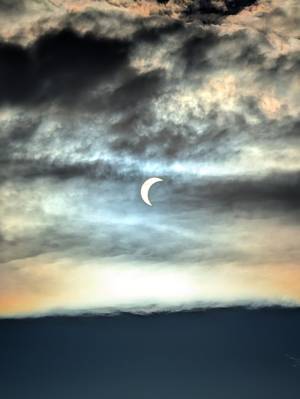 A luminous crescent sun glows near the centre of a dark, dramatic sky filled with heavy, sweeping cloud formations. Soft pastel tones of warm gold, light orange, and pale cyan bleed through the hazy cloud layers beneath the crescent, contrasting sharply with the deep dark blues and shadowy charcoals framing the top and bottom of the scene.
