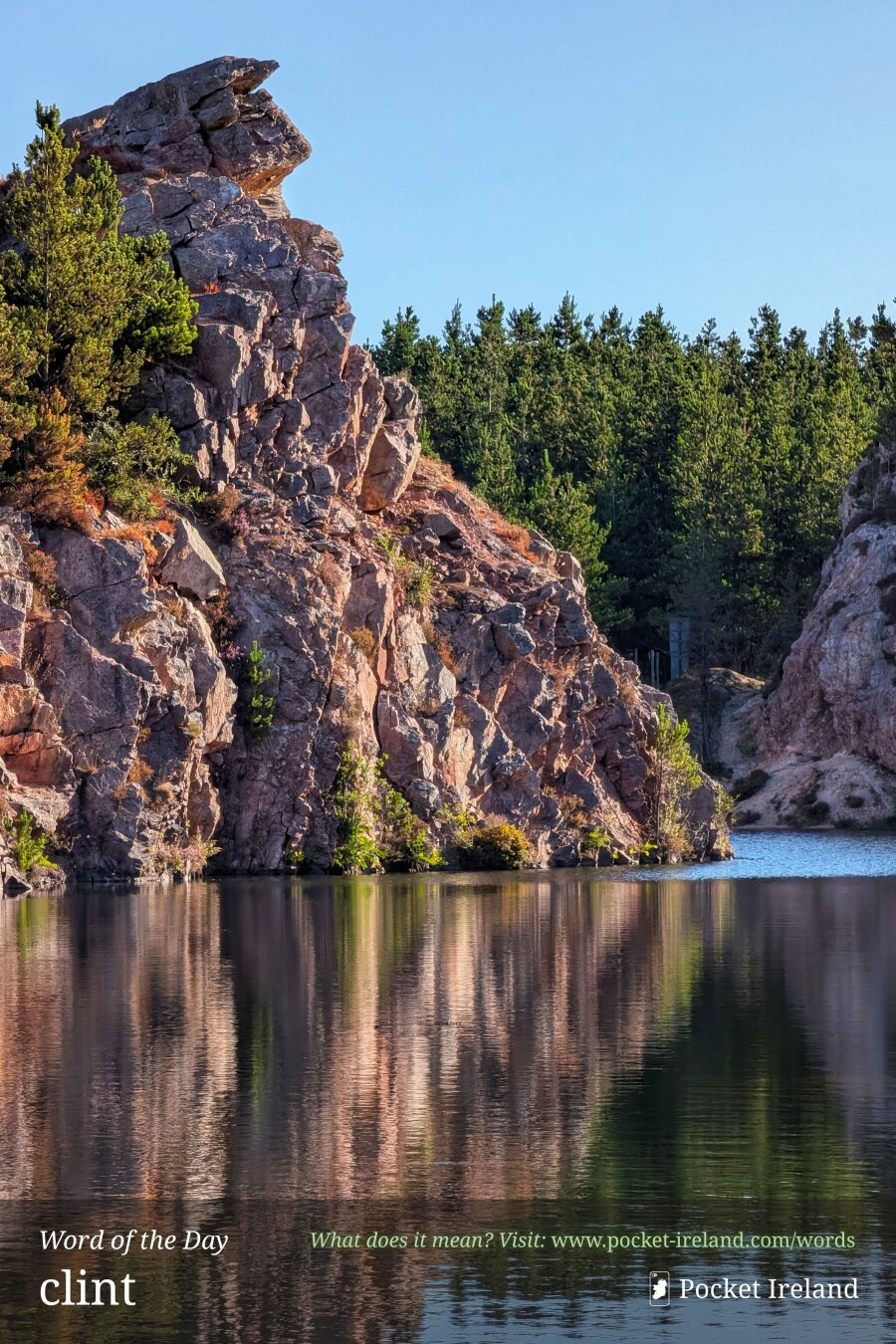A dramatic, sunlit rocky crag of fractured pink and grey stone rises sharply above a calm, glassy lake, casting rippling vertical reflections across the dark water. Deep green conifer forest blankets the crest and background under a clear pale blue sky.