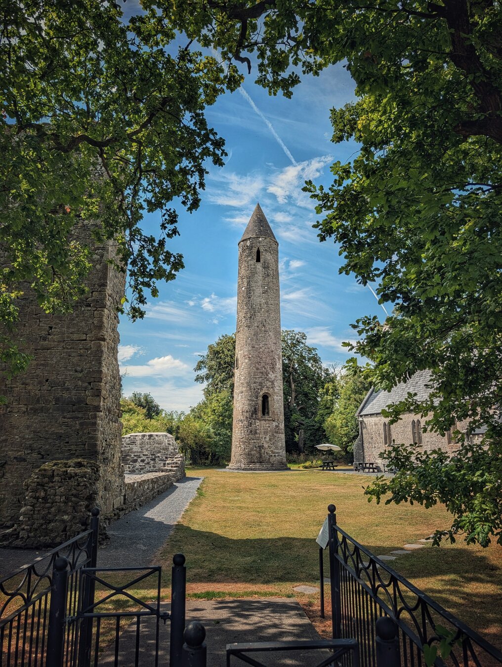 A vertical, low-angle photo capturing a tall grey stone round tower framed by leafy green branches above. The tower rises into a bright blue sky with light white clouds. To the left sit the ruins of an old stone building, while a small grey church stands on the right. In the foreground, a dark metal fence opens onto a gravel path across a sunlit green lawn, leading towards the central tower.