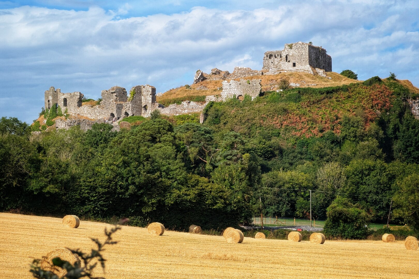 A wide landscape photograph featuring a golden, freshly harvested field dotted with round hay bales across the foreground. In the midground, a steep hill covered in lush green trees and foliage rises toward the rugged stone ruins of an ancient hilltop fortress. Above, soft white clouds drift across a vibrant blue summer sky, creating a bright, peaceful, and historic countryside scene.