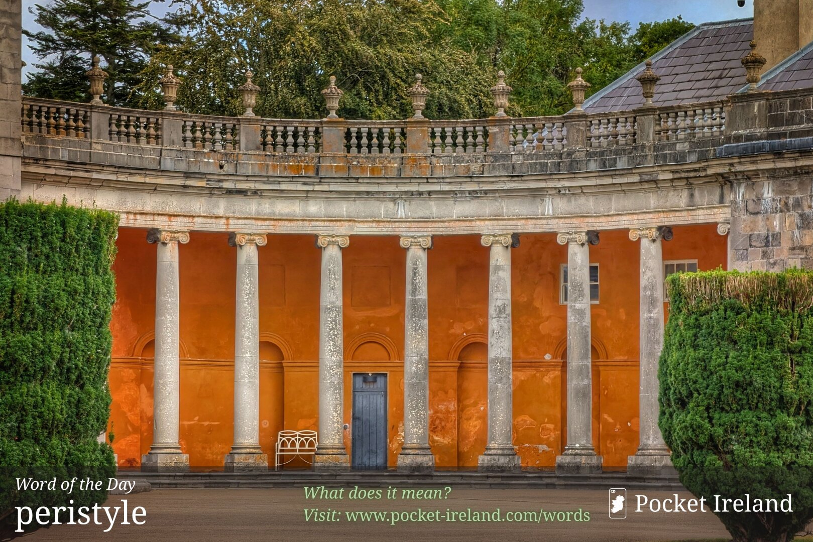 A curved neoclassical stone colonnade with weathered grey Ionic pillars set against a vibrant terracotta-orange wall. An ornate stone balustrade with decorative urn-shaped finials tops the structure under an overcast sky. In the shadowy alcove behind the pillars, a dark wooden doorway sits adjacent to a white wrought-iron bench. Dense green manicured hedges frame the foreground on both sides, while lush tree foliage rises softly in the background.