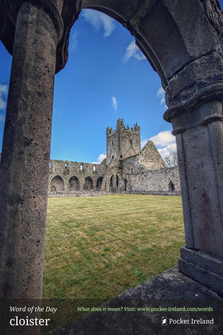 Framed by dark, weathered stone pillars and a arched doorway in the foreground, the image opens onto a sunny, grassy courtyard surrounded by ancient stone monastery ruins. A central crenellated square tower stands tall against a vibrant blue sky dotted with soft white clouds, flanked by stone walls with arched cloister openings and ruined gables under bright day lighting.