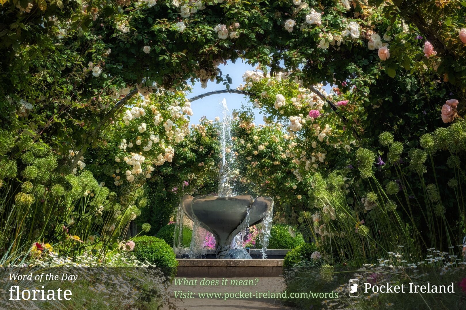 A light-dappled gravel path leads toward a decorative metal water fountain spraying water into a bright sky. The pathway is framed by lush garden arches cascading with cream and soft pink roses. White daisies and tall round seed heads line the borders below, while sunlight illuminates the cascading water, green foliage, and vibrant blossoms, creating an enchanting, romantic garden setting.