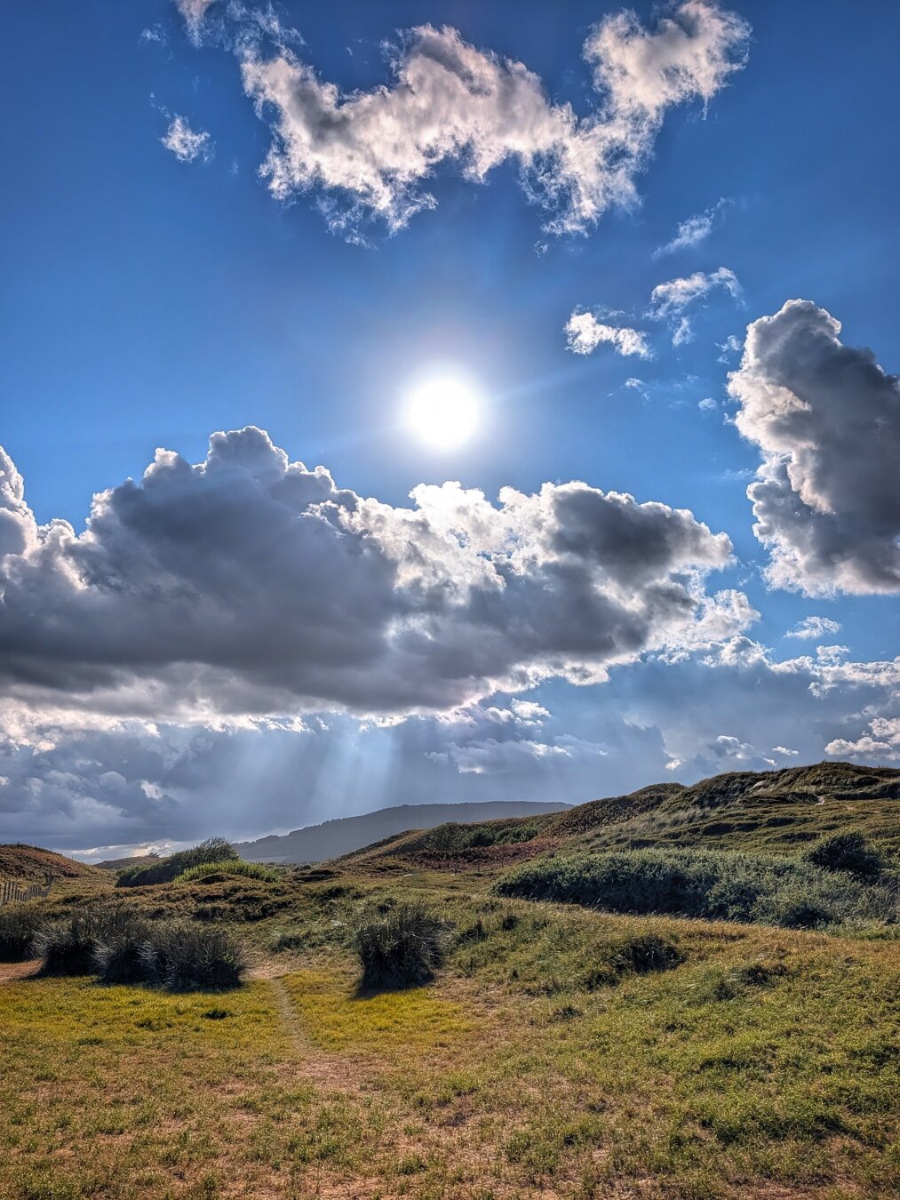 A vertical, eye-level photograph of rolling sand dunes covered in green and brown grasses under a dramatic blue sky. Near the top centre, the bright sun shines through a gap in thick, dark grey and white clouds, sending visible beams of light down towards the terrain. Low, dark hills line the horizon in the background. In the foreground, sandy paths wind between patches of grass and shrubs, creating a textured coastal landscape bathed in bright, natural sunlight and deep shadows.