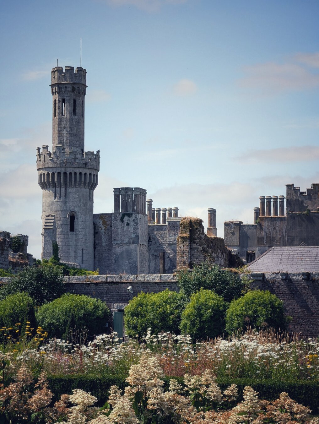 A tall, pale grey Gothic Revival stone turret and ruined castle parapets rise against a pale blue sky with light clouds. In the foreground, cream-coloured astilbe and mixed summer meadow flowers bloom in a lush walled garden, separated from the weathered stone battlements and brickwork by a row of neatly clipped green shrubs.