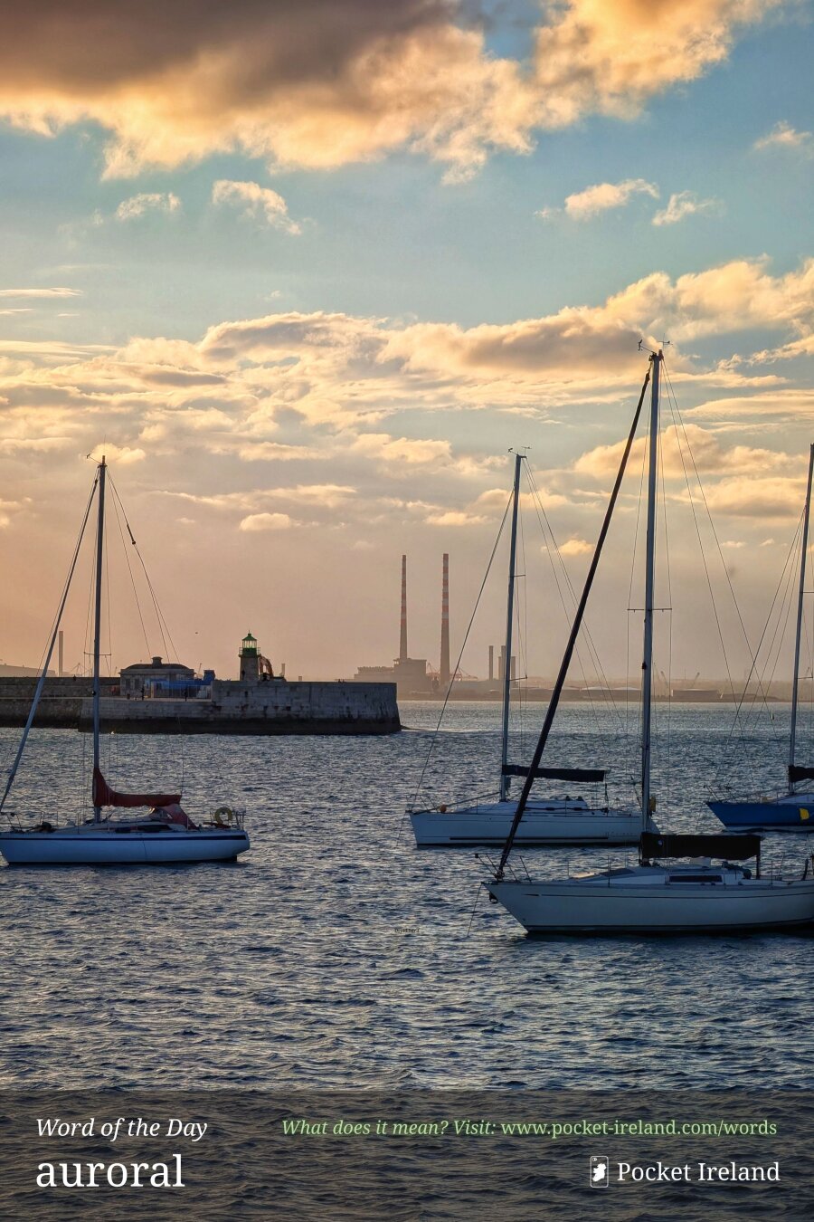 Several yachts are moored on calm water at the edge of a harbour under a cloudy sky. One yacht is prominent in the foreground, facing right, with another to its left. Behind them, a long concrete breakwater extends from the left, ending at a small green-roofed building and a short lighthouse structure. Far in the distance on the hazy horizon, two very tall, red-and-white industrial chimneys are visible. The setting sun illuminates the tops of the clouds in shades of peach, orange, and pale yellow, contrasting with the blue and grey of the water.
