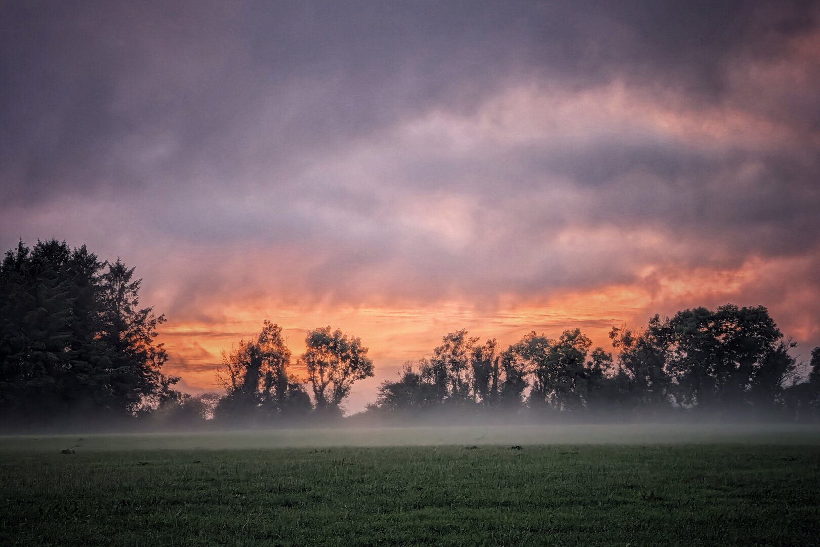 A rural landscape photographed at sunset on a calm evening. A flat green field in the foreground is blanketed by a low layer of white ground mist. In the middle ground, a silhouette of mature deciduous and evergreen trees lines the boundary. Behind the tree line, the horizon glows with intense bands of fiery orange and soft amber light. Above the glow, heavy, textured clouds in deep shades of purple and slate grey fill the rest of the sky. The scene conveys a quiet, cool, and atmospheric mood as dusk settles over the countryside.