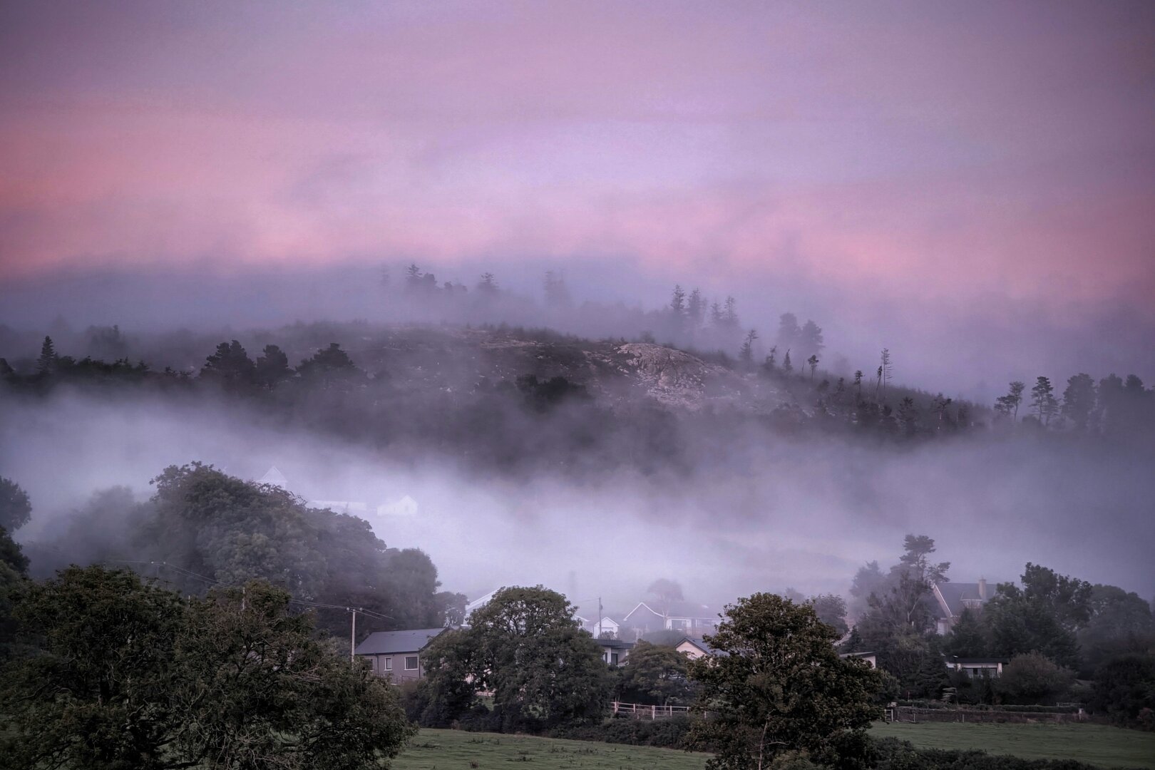 A landscape photograph taken at dusk showing a green hill rising above a thick band of white mist. In the foreground, dark silhouettes of tree canopies and green fields surround a few small houses partially veiled by fog. A dense ribbon of mist cuts horizontally across the middle of the frame, softening the lower hillside. Above the mist, the rocky, wooded crest of the hill stands clear beneath a smooth, tranquil sky glowing in shades of dusky rose, lilac, and soft mauve.