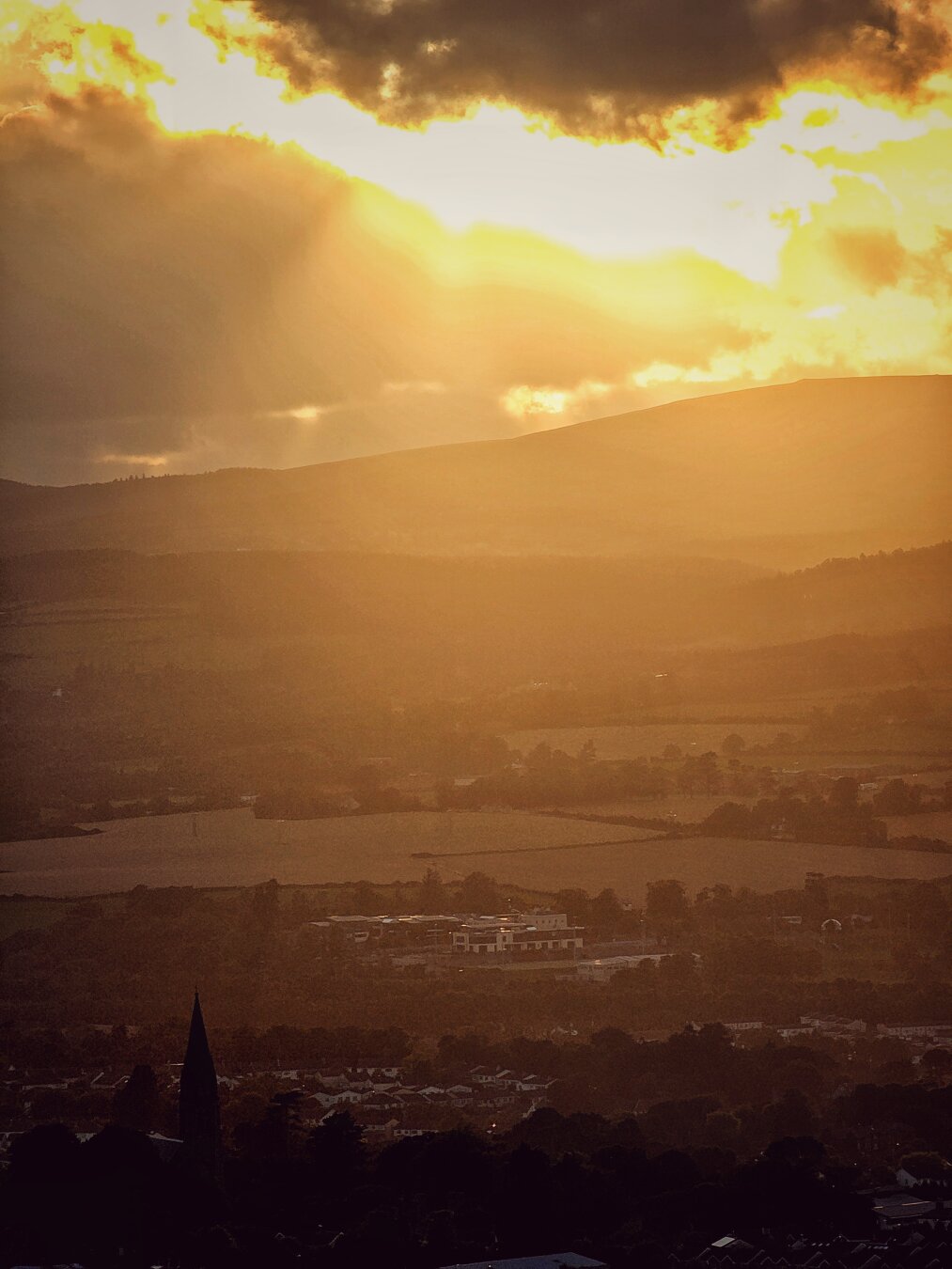A vertical, atmospheric landscape photo looking down from a high vantage point over a misty valley and distant rolling hills at sunset. The sky is filled with dark, stormy clouds at the top, which part to reveal bright, golden sunlight. Broad sunbeams stream down across the hazy hills and farmland below. In the shadowed lower foreground, tree canopies and a dark, slender church spire rise gently against the warm, amber glow of the valley floor.