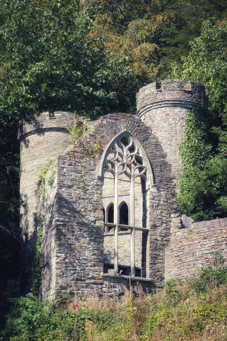 A vertical eye-level view of an old grey stone folly built into a steep, wooded hillside. Two rounded towers with castellated tops frame a tall Gothic arch filled with delicate, pale stone tracery. Sunlight catches the weathered masonry, while climbing green ivy and woodland foliage wrap around the structure, creating an atmospheric, fairy-tale mood.