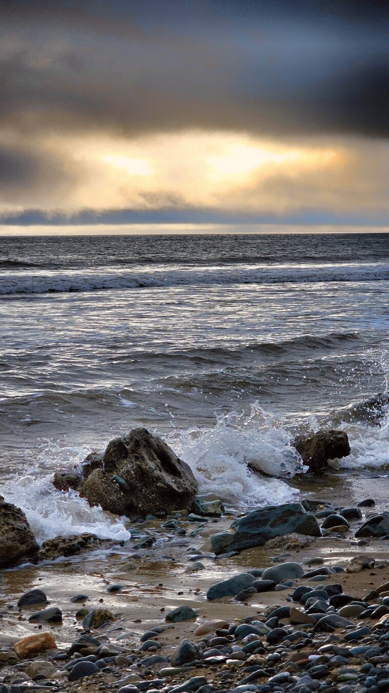 A vertical, eye-level photograph of a rugged Irish shoreline. In the foreground, white foam splashes over dark, wet rocks and smooth pebbles on a sandy beach. The midground shows dark, rippling ocean waves stretching toward the horizon. Above the sea, dramatic dark storm clouds hang in the upper sky, broken horizontally across the centre by a warm, golden glowing band of sunlight illuminating the sea spray. The overall mood is cold, dramatic, and atmospheric.