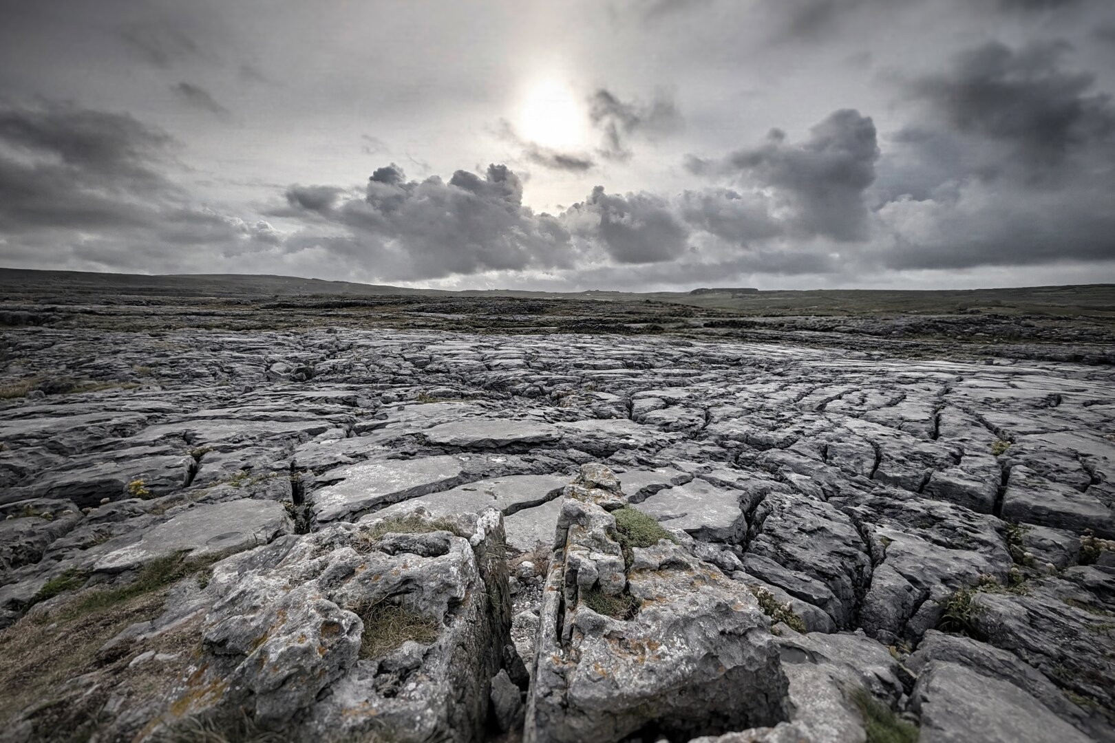 A wide landscape photograph of an expansive limestone pavement stretching towards a distant low horizon under a dramatic, overcast sky. The pale sun glows faintly behind layers of dark, textured storm clouds. In the foreground, weathered grey limestone blocks feature deep, dark crevices, fissures, and small patches of green moss and dry lichen. The perspective emphasizes the vast, cracked geometric stone field and the sombre, cool grey and muted earth tones across the wild terrain.