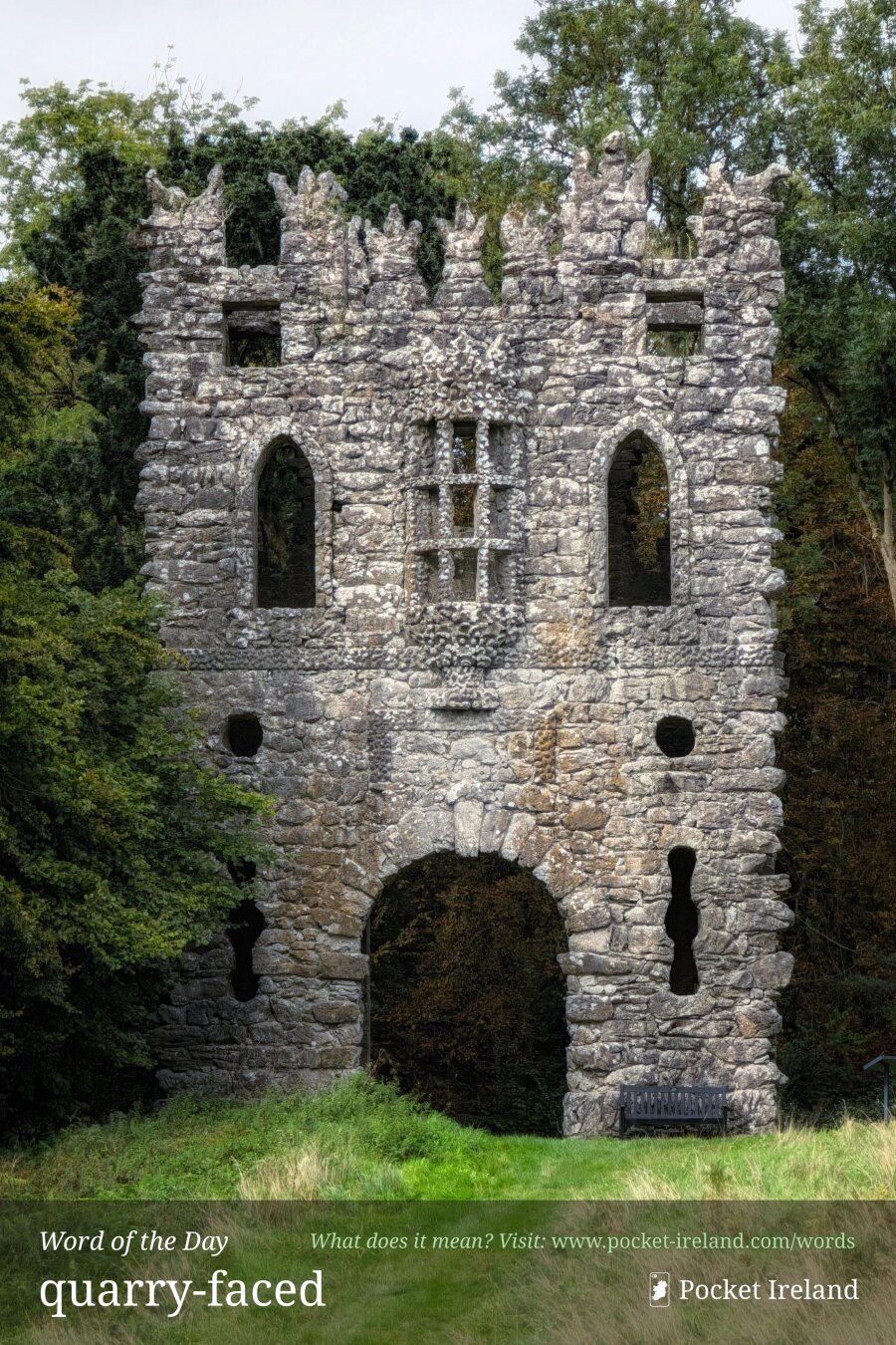 An imposing, weathered stone gothic folly tower rises from a grassy slope, flanked by dense green woodland. Built from rough, uncoursed grey fieldstones, the structure features a large central arched gateway at ground level, narrow vertical arrow slits, and arched lancet window openings above. The parapet is adorned with jagged, decorative crenellations. Diffuse, soft daylight illuminates the tactile masonry, evoking an ancient, secluded atmosphere.