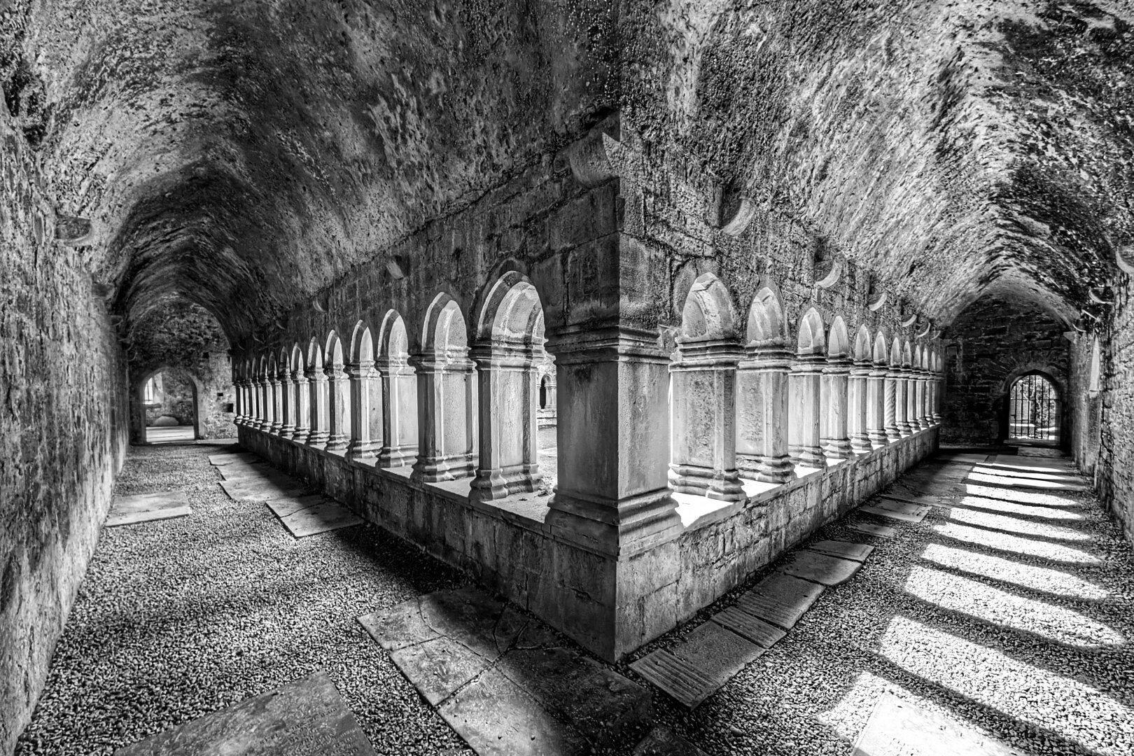A wide black and white photograph captured from the corner angle inside the covered stone cloister of an ancient abbey. An unbroken sequence of pointed medieval stone arches supported by short paired columns runs along two perpendicular sides of a gravel walkway. Sunlight streams low through the arched openings, casting sharp, rhythmic rectangular bars of light and deep diagonal shadows across the gravel floor and sunken stone slabs. Overhead, a curved, coarse barrel-vaulted ceiling shows weathered masonry texture leading towards arched doorways at each corridor end.