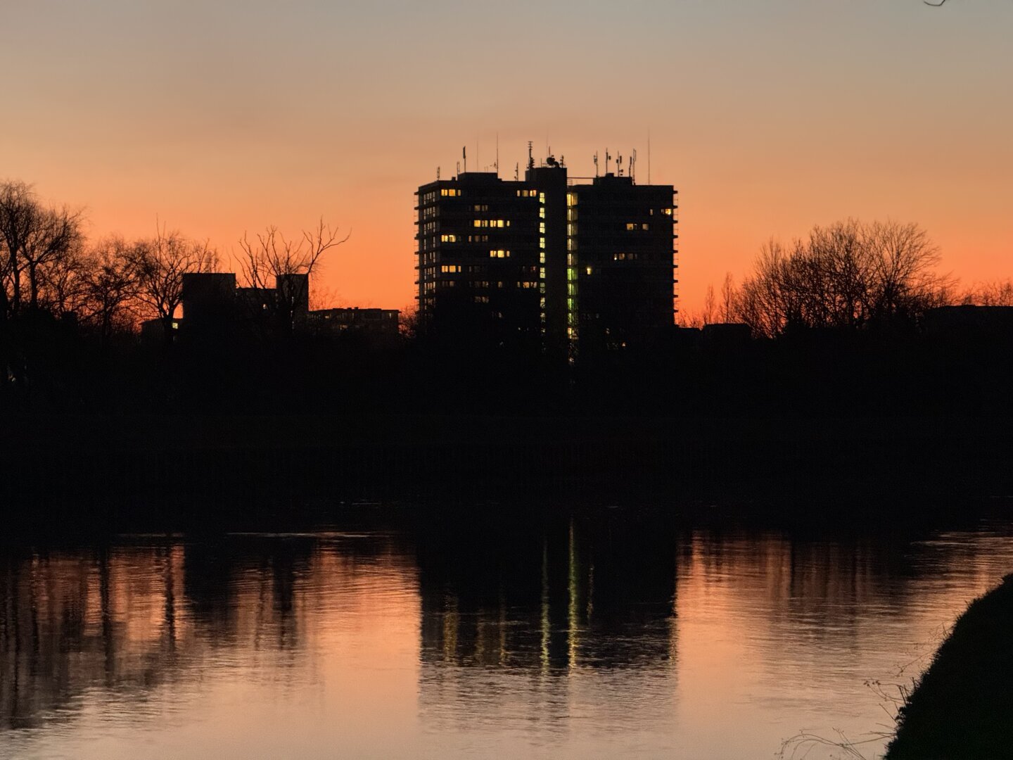 A stunning sunset over the Neckar River in Heidelberg, with the silhouette of a high-rise building in Wieblingen reflecting in the calm waters. The warm hues of the evening sky create a breathtaking contrast against the dark outlines of trees and architecture.