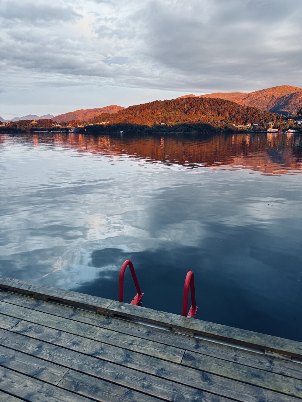 This image depicts a serene outdoor scene at dusk or dawn, featuring a calm body of water bordered by a wooden dock and a distant landscape.
Key elements of the image:
 * Water: The dominant feature is the water, which appears smooth with gentle ripples. It reflects the colors of the sky and the surrounding landscape, creating a sense of depth and tranquility. The reflection of the sky suggests a partly cloudy day, with lighter tones mirroring potential sunlight or breaks in the cloud cover.
 * Dock: A wooden dock occupies the lower portion of the image, extending into the water. The dock's weathered planks show texture and signs of outdoor exposure. A prominent feature on the dock is a bright red ladder descending into the water, suggesting easy access for swimming or boating.
 * Landscape: In the distance, across the water, a hilly landscape is visible. The hillsides are covered with trees, transitioning from lush green at the base to a warm, sun-kissed orange-brown hue towards the p