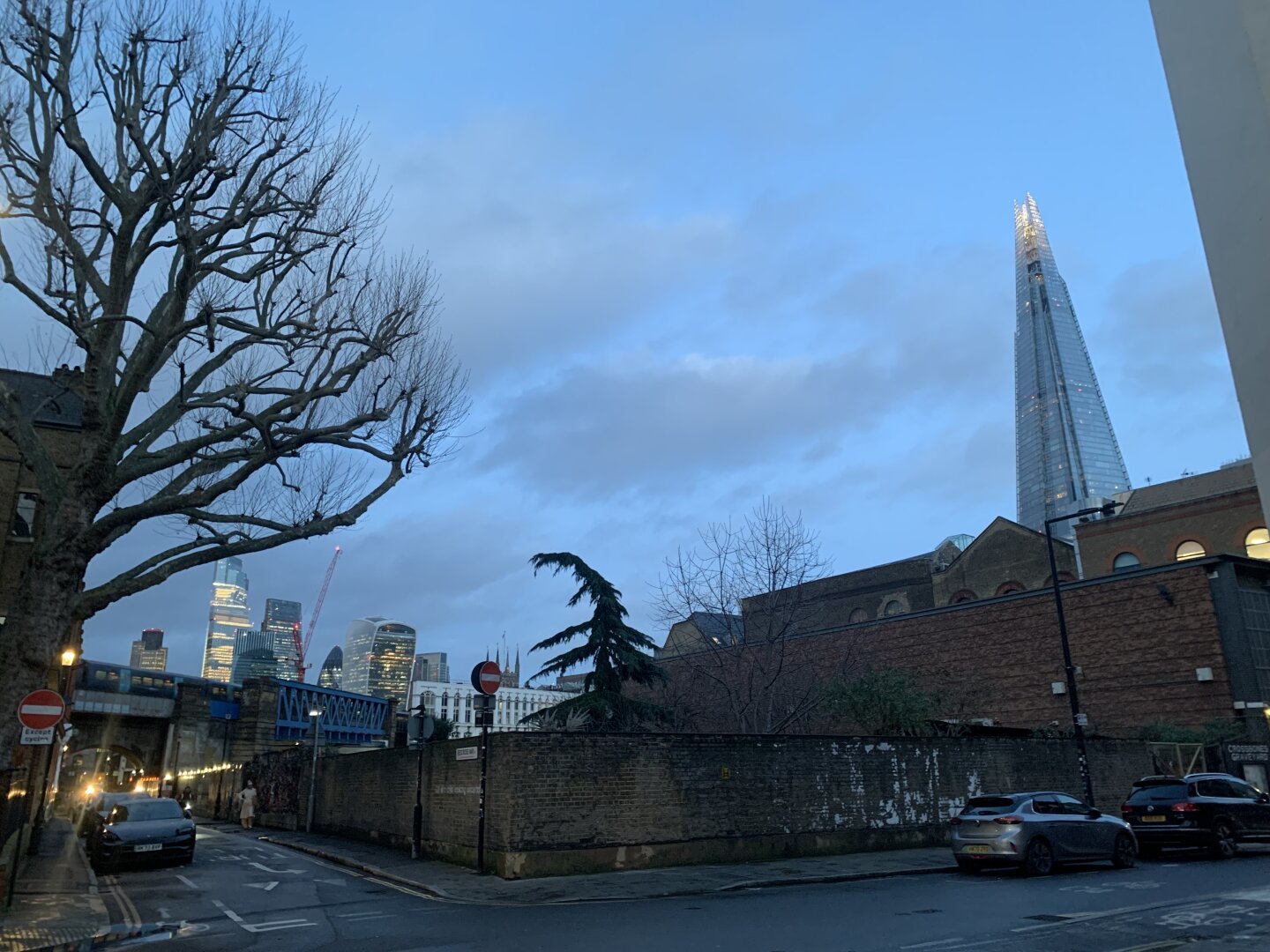 A pic I took last night of a crossroads with trees and a garden nearby and tall city buildings in the background. The general landscape is Victorian warehouse like. It is dusk and many lights are on in the glass towerblocks.