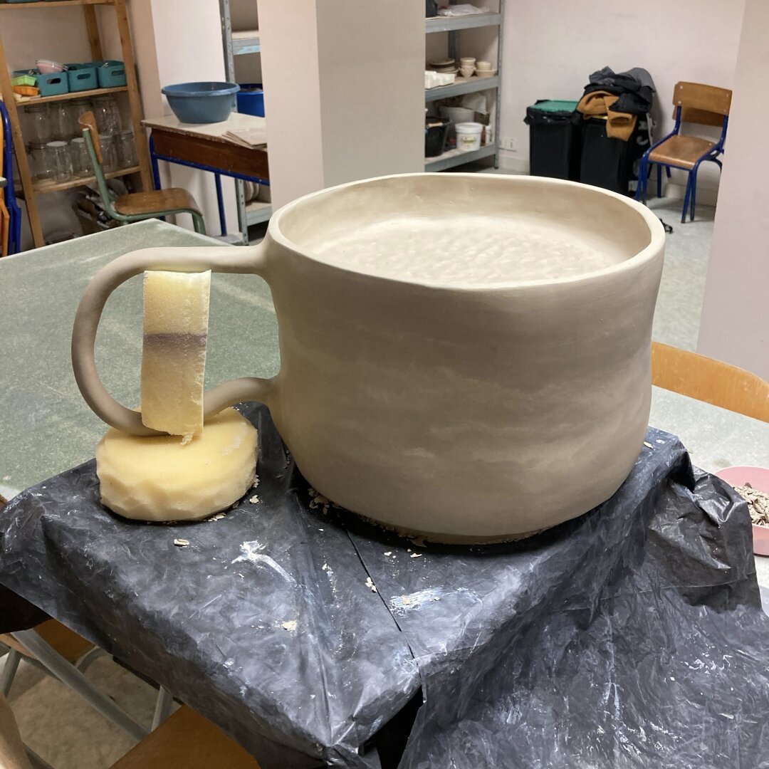 Photo d’une tasse surdimensionnée réalisée en céramique, dans un atelier (l’anse est en train de sécher et des petits morceaux de mousses sont la pour soutenir le temps du séchage)