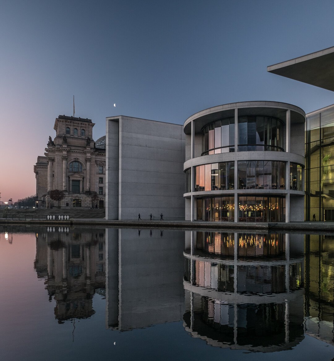 The image shows the Berlin government district at dusk. On the left is the Reichstag, a historic building with a dome and ornate facades. On the right is the modern Paul-Löbe-Haus, featuring a round glass façade illuminated with warm interior lights. Between the two buildings stands a tall, rectangular concrete structure that is part of the architectural ensemble. The entire scene is reflected clearly on the calm surface of a canal in front of the buildings. In the sky, a crescent moon is visible, with colors transitioning from pink to deep blue. Three people appear as small silhouettes along the canal.