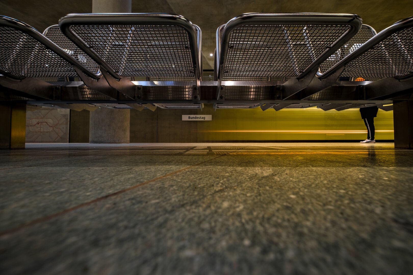 The image captures a subway station scene from a low-to-the-ground perspective. In the foreground, several metal benches with a grid-like structure are visible from below. Through the gap between the seats, a yellowish blurred subway train is visible in the background, indicating motion. On the right side of the image, the legs of a person wearing dark clothing and white sneakers can be seen. A sign on the wall in the background reads “Bundestag,” indicating that this is a station in Berlin. The floor consists of large, greenish stone tiles with a slightly reflective surface.