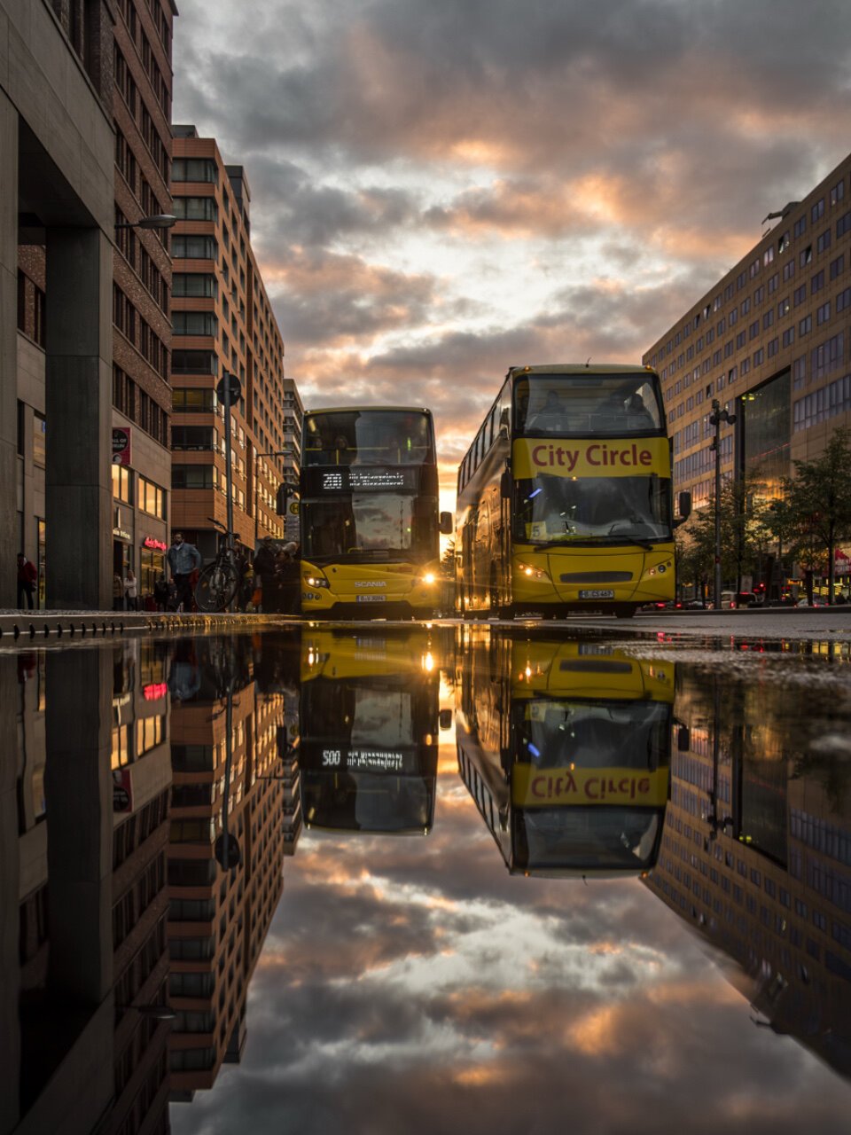 The image shows two yellow double-decker buses standing side by side on a street. The bus on the left displays route “200,” while the bus on the right features the text “City Circle,” indicating a city tour. The scene is dominated by an impressive evening atmosphere with dramatic clouds and warm, golden light. In the foreground, a large water surface reflects the buses, buildings, and sky. The surrounding buildings have modern facades, some made of brick and glass, emphasizing the urban character of the scene. People and bicycles are subtly visible in the background.