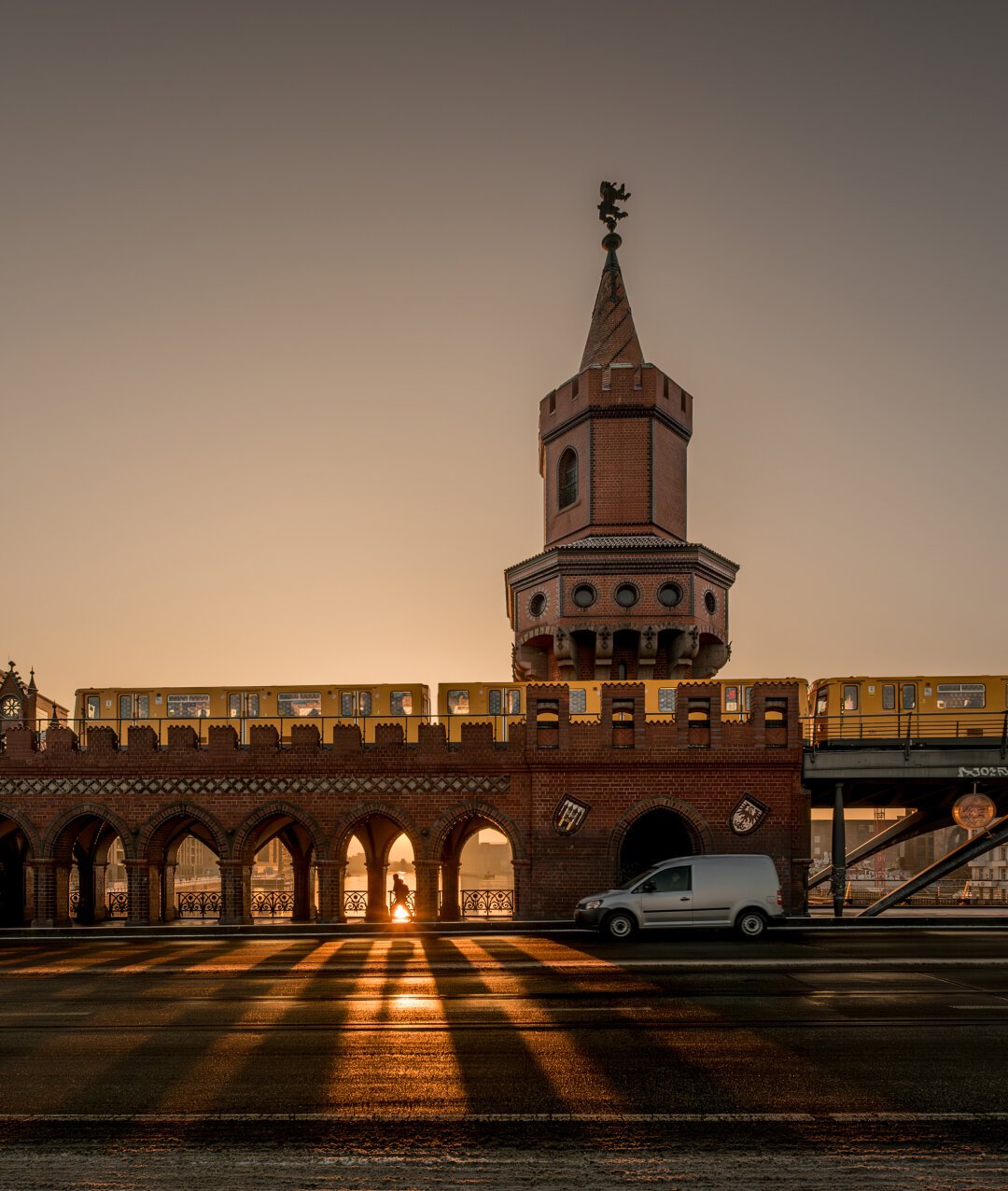 The image shows the Oberbaum Bridge in Berlin at sunrise. In the upper section, a yellow subway train of the Berliner Verkehrsbetriebe (BVG) crosses the bridge. The iconic brick tower of the bridge with its pointed spire rises prominently into the sky. In the lower section, the arches of the bridge allow the warm light of the rising sun to shine through, casting long shadows across the street. On the road in the foreground, a white delivery van is driving from right to left. The sky is clear and painted in soft yellow and brown tones, highlighting the tranquil morning atmosphere.