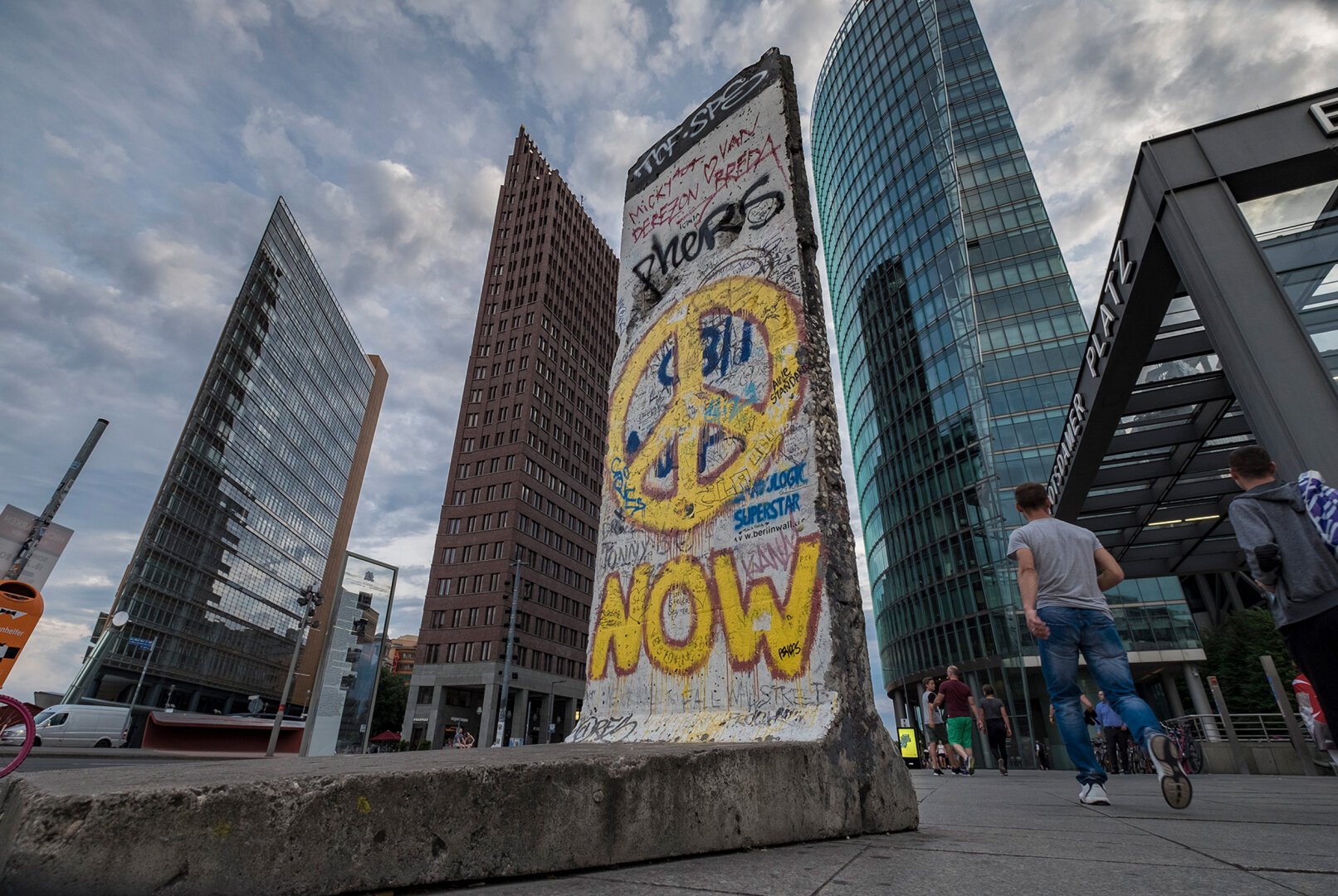 The image shows a segment of the Berlin Wall, painted with graffiti including a large yellow peace symbol and the word “NOW.” The wall piece stands upright on a public square at Potsdamer Platz in Berlin. In the background, three modern buildings are visible: on the left, a glass building with a pointed shape; in the center, a reddish-brown high-rise; and on the right, a rounded glass skyscraper. On the far right, part of a building entrance with the sign “POTSDAMER PLATZ” is visible. Several people are walking or standing on the square, including a man in a gray T-shirt and blue jeans in the foreground. The sky is cloudy.