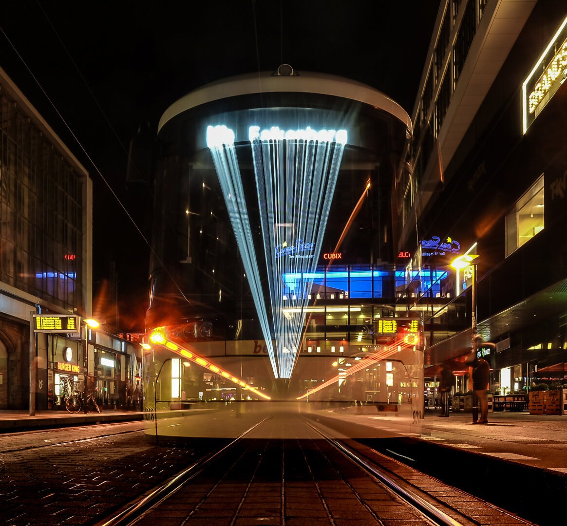 The image shows a tram at a stop during the night. The tram dominates the center of the frame. Through long exposure, bright light trails appear at the front of the tram, fanning out from the upper middle downward. On both sides of the tram, illuminated buildings are visible. To the left, shop windows and a “Burger King” logo can be seen. To the right, colorful neon signs, including the “Cubix” cinema, light up the area. The ground is made of cobblestones, and tram tracks run diagonally through the image. In the background, stop displays and a few pedestrians are visible.