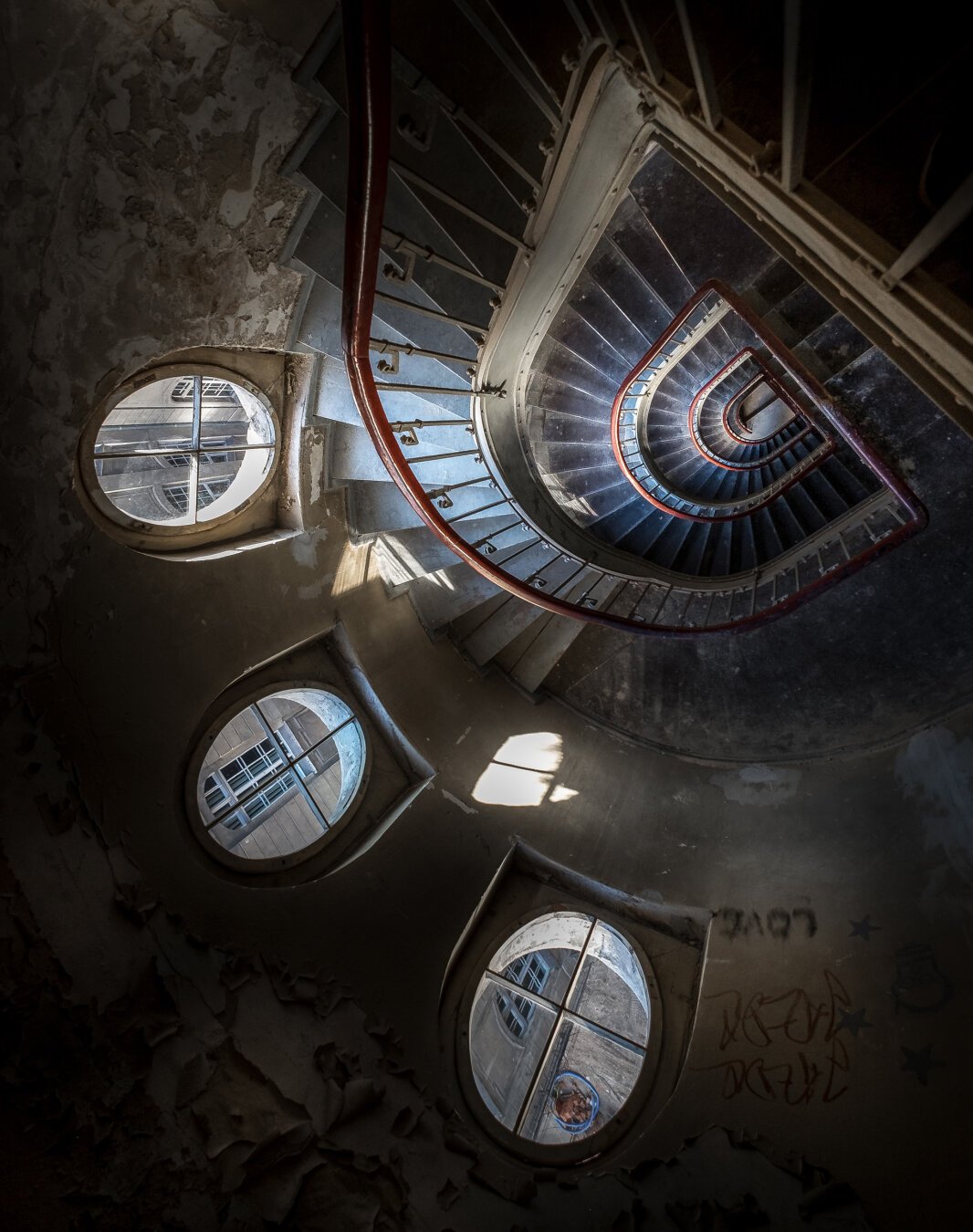 The image shows a top-down view of a spiral staircase with worn and weathered walls. The staircase curves elliptically downward and is surrounded by a red handrail. The steps are in shades of dark blue and gray. Four round windows in the walls let daylight enter, casting shadows onto the uneven, peeling wall paint. Graffiti and scribbles can be seen on the lower walls. Through the windows, the facade of a neighboring building is visible. The overall atmosphere of the space feels abandoned and dilapidated.