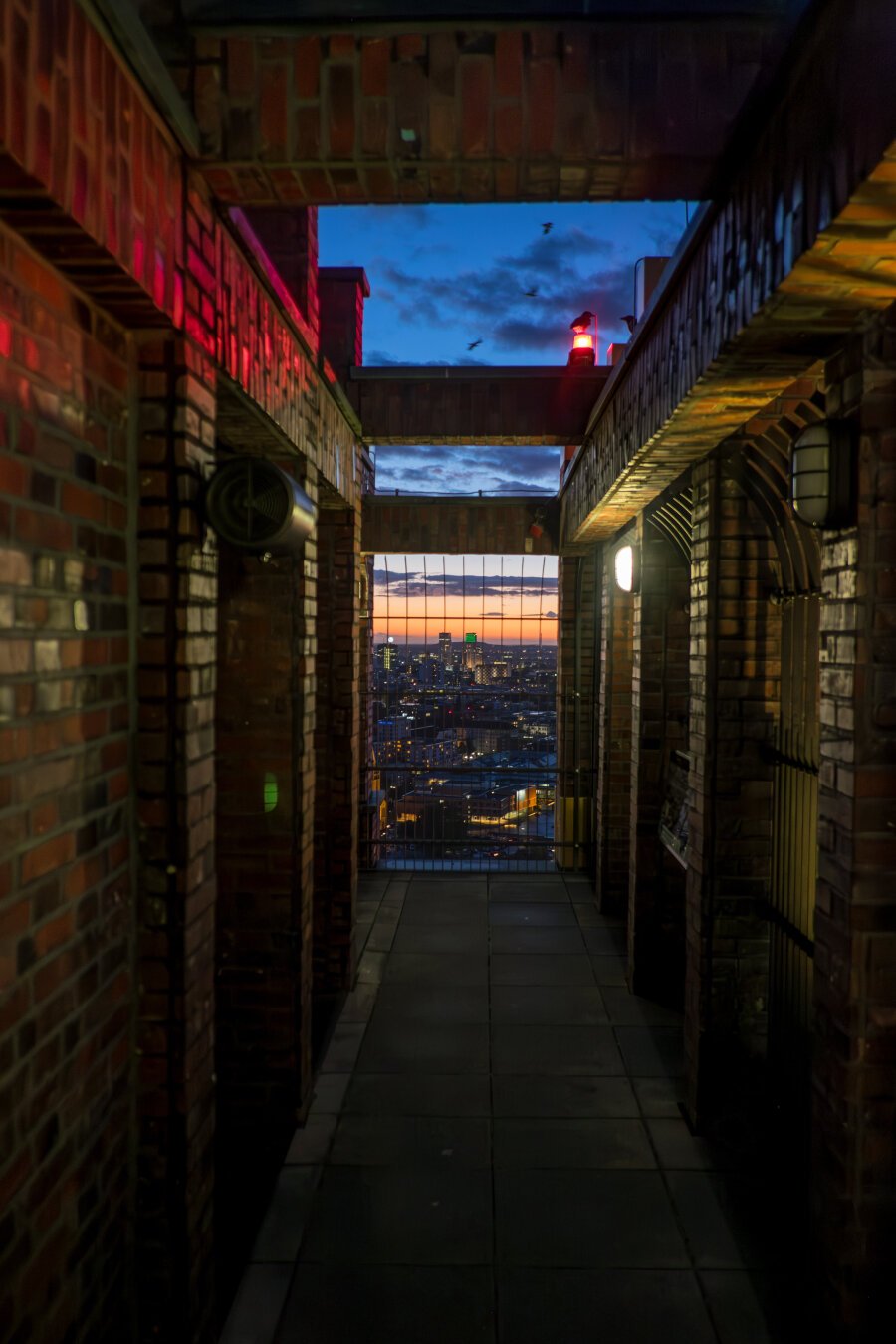 The image shows a narrow, covered passageway with brick walls leading to a gated view of a cityscape at dusk. The bricks reflect warm light, including red and yellow tones from artificial light sources installed in the corridor. At the end of the passage, there is an expansive view of the skyline, illuminated by the last streaks of sunset and the city’s early lights. At the top of the corridor, a red warning light is visible, and birds are flying against the blue evening sky.