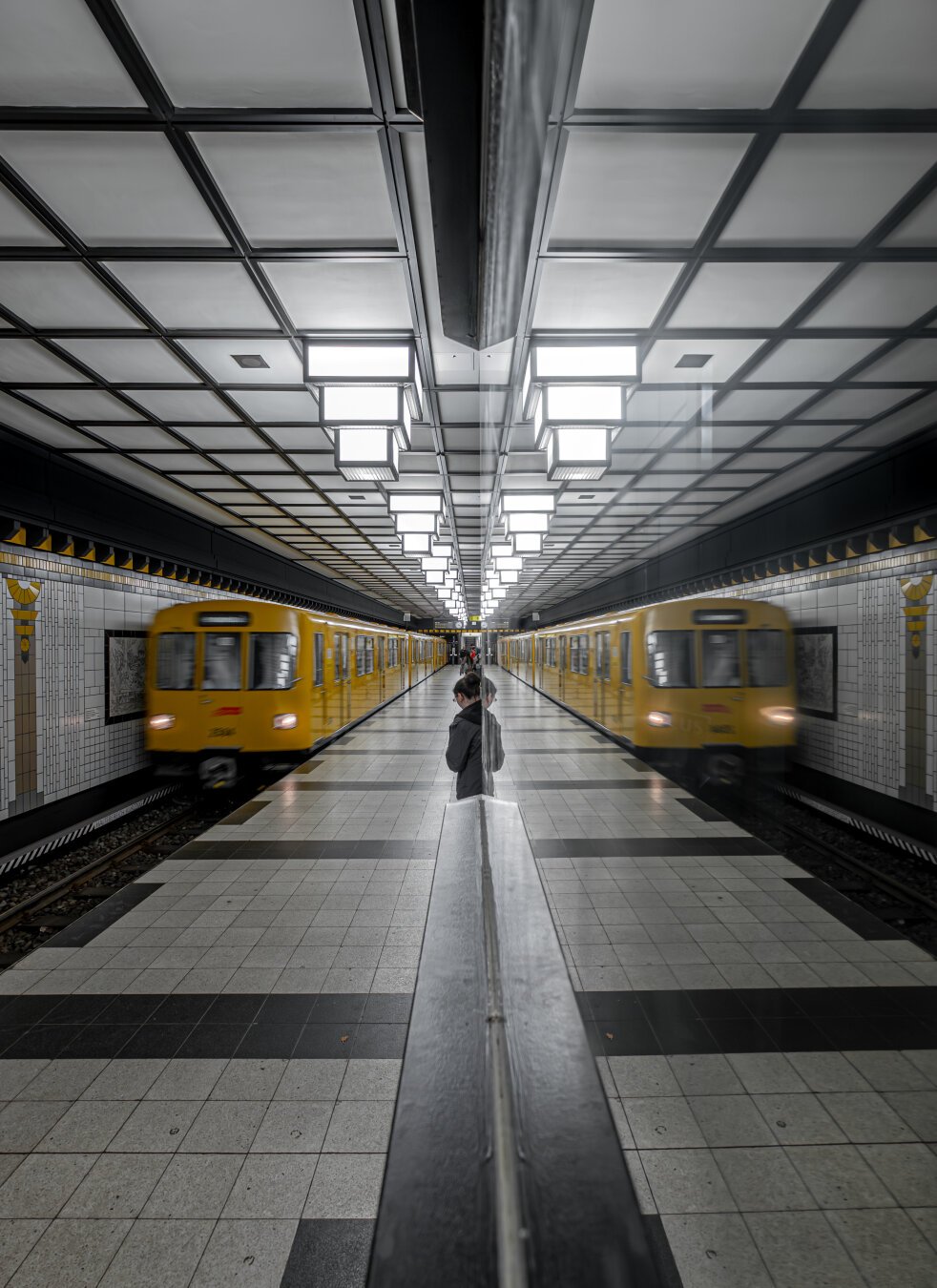 The image shows a subway station with a symmetrical design. Two yellow subway trains are arriving simultaneously on opposite tracks. In the center of the image is a railing with a reflective surface, mirroring the station and a waiting person. The person is standing on the platform, looking toward one of the arriving trains. The station’s ceiling features a grid pattern of illuminated squares, providing even lighting throughout. The walls are tiled in white, with yellow and black accents, and the floor is decorated with a geometric pattern of black and white tiles.