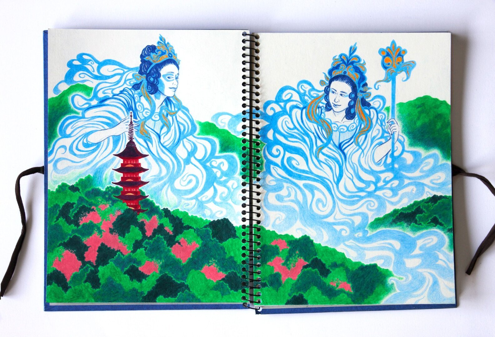 This is an illustration drawn with colored pencils in an open sketchbook. Two delicate female figures appear as swirling cloud-like forms in shades of blue. One holds an ornate staff, while the other seems to be observing the red pagoda below. The landscape features rolling hills covered in dense vegetation, with hints of pink contrasting against deep greens. Their fluid shapes stretch above the mountains and weave through the valleys, following the curves of the terrain.