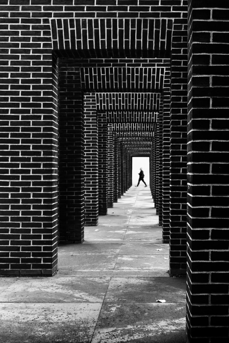 die silhouette einer laufenden frau in der bildtiefe, wird von einer aneinanderreihung dunkler wandscheiben aus kleinen ziegelsteinen eingerahmt. man blickt wie durch einen tunnel auf die kleine silhouette. der mauerrahmen ist durch die dunklen ziegelsteine und weiße fugen sehr kontrastreich.