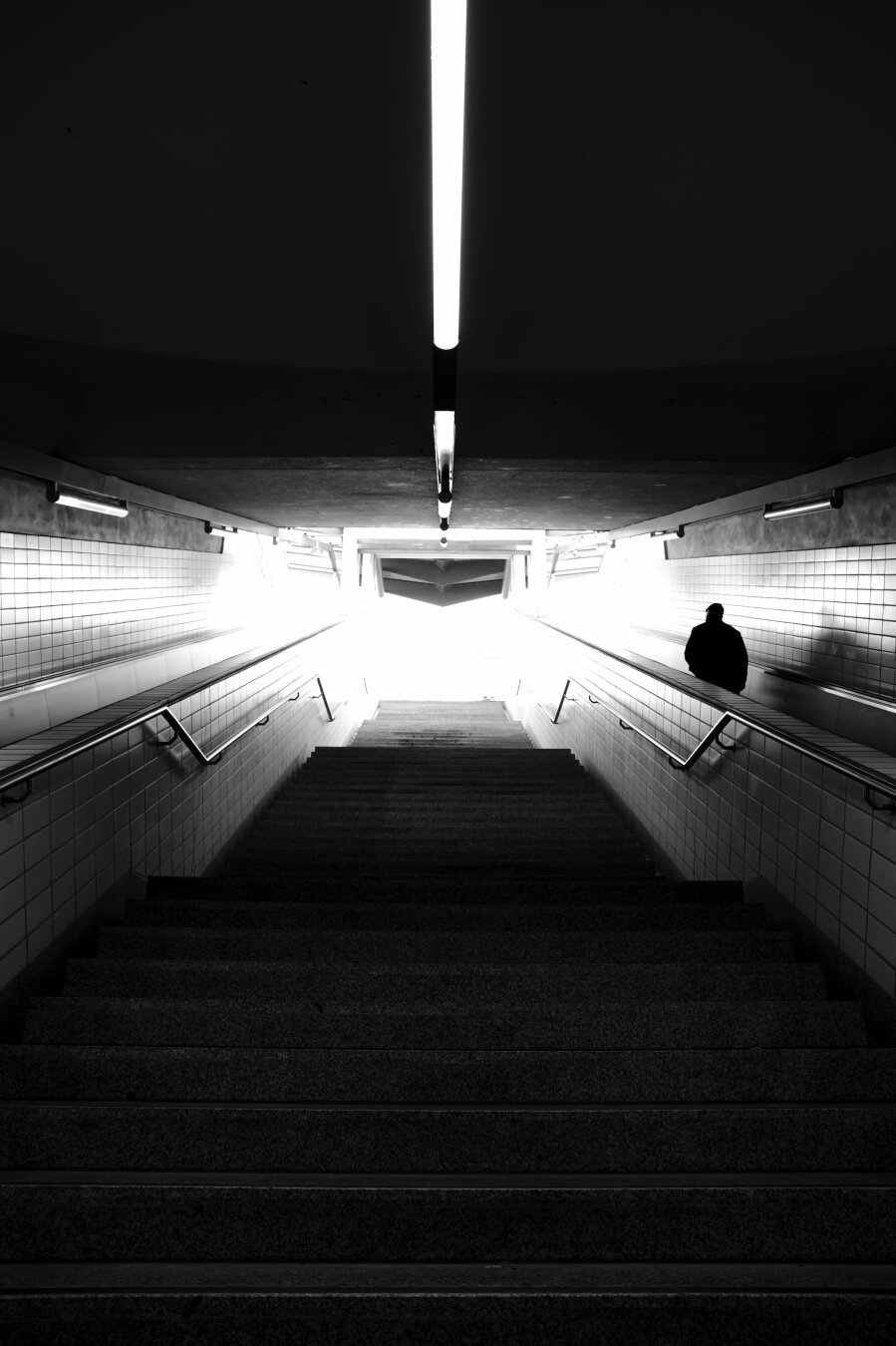 schwarzweißbild. treppenaufgang einer u-bahnstation in frankfurt. von unten nach oben fotografiert. unten dunkel. oben hell. gegenlicht. starke kontraste. passant als dunkle silhouette auf rolltreppe am rechten bildrand. linien und kontrastreiche flächen erinnern mich an geometrie des fluxkompensators aus dem film zurück in die zukunft.