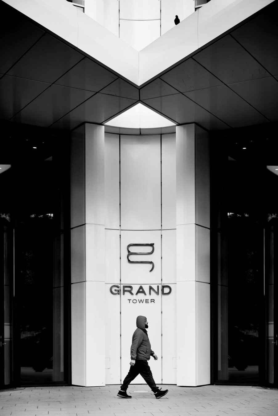 a darkly dressed man with a hood and a striking gait is photographed in the middle in front of the symmetrical entrance area of a high-rise apartment building with a white facade. the photo is very clean and symmetrical overall. this tidy look is cheekily ignored and loosened up by a dark dove sitting on the front roof.