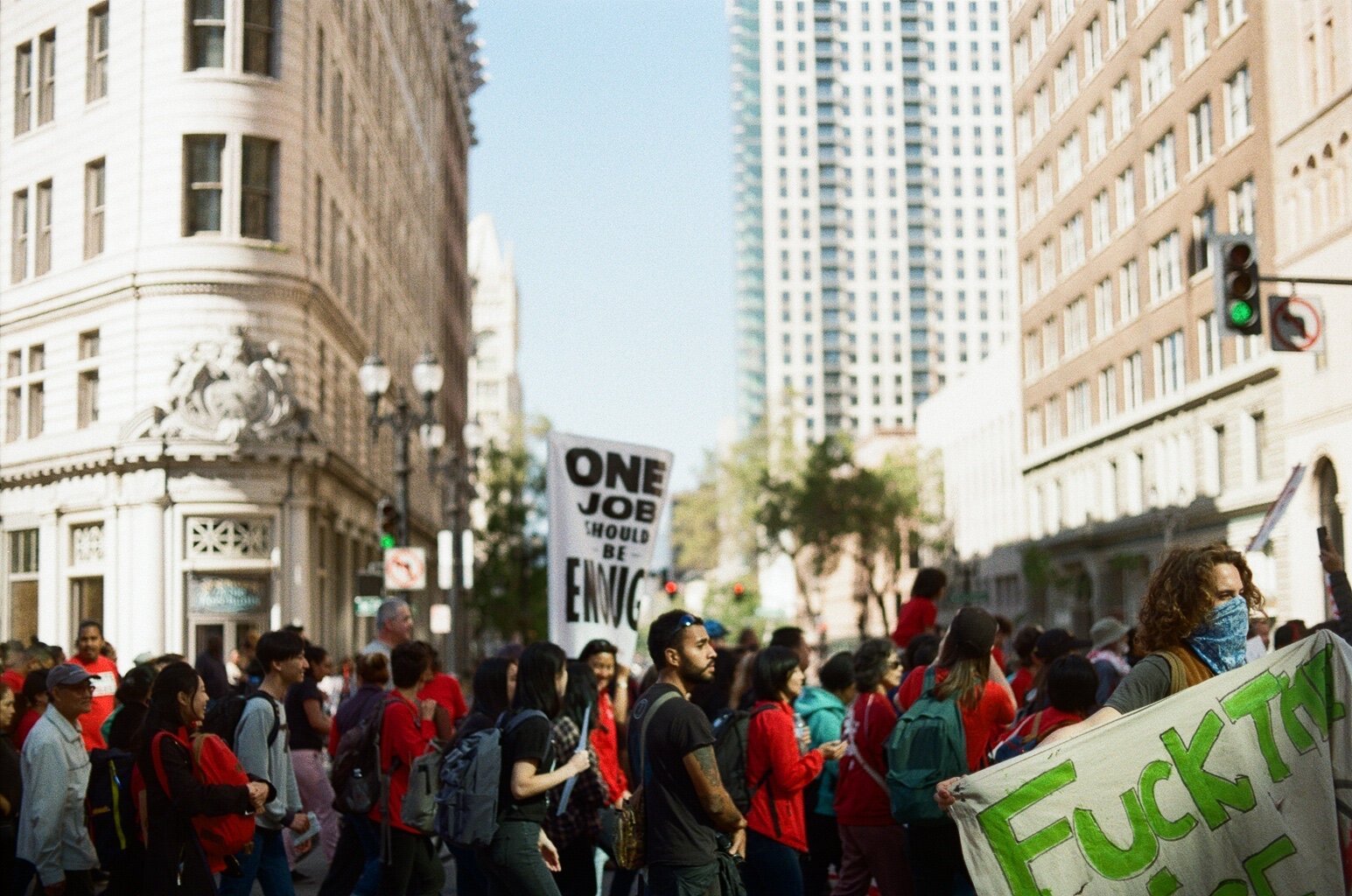A protest crowd of City of Oakland union workers marched dow 14th street at broadway. A sign in the background declares “one job should be enough”
