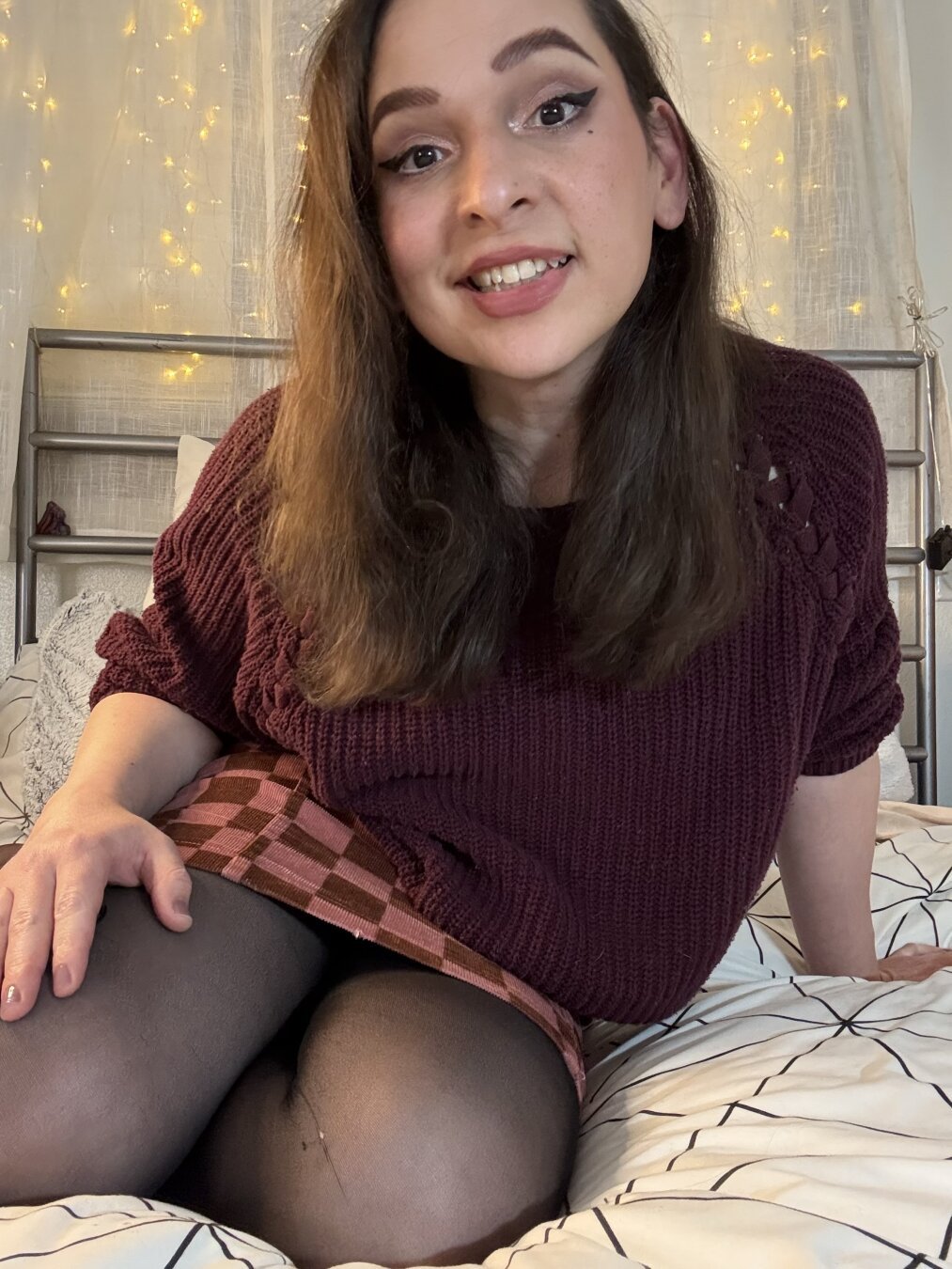 A portrait of a cute trans woman (Bijou), smiling while kneeling on a bed, and making eye contact. She has long dark brown hair, and is wearing eyeshadow, lipstick, winged eyeliner, a maroon colored sweater and red checker skirt, and black nylons. Room interior.