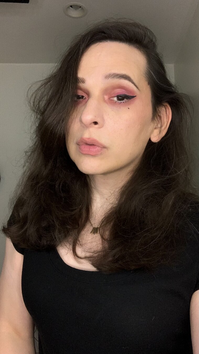 A selfie of a cute trans woman (Bijou) making eye contact. Neutral expression. She has wavy long dark brown hair, and is wearing red eyeshadow, winged eyeliner, a black dress, and a necklace. Room interior.