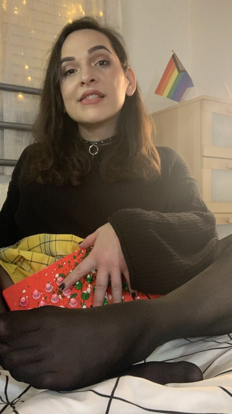 A portrait of a cute trans woman (Bijou), smiling while sitting on a bed crisscross applesauce, and making eye contact, holding a  splatter painted midi controller between her legs. She has long dark brown hair, and is wearing a black sweater, yellow plaid skirt, a choker necklace, and black nylons. Room interior. Lgbtqia+ flag in the background.