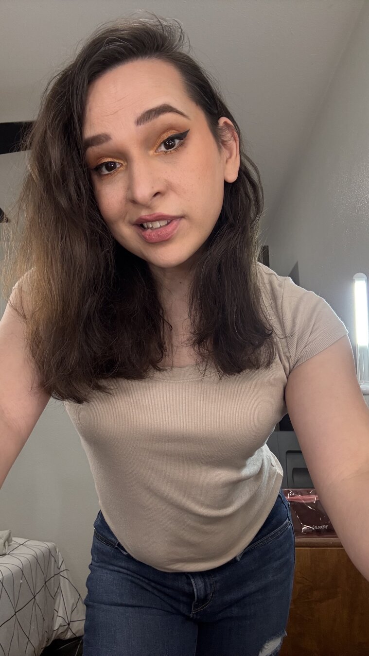 A selfie of a cute trans woman (Bijou), smiling coyly while leaning towards the camera, and making eye contact. She has long dark brown hair, and is wearing golden eyeshadow, neutral lipstick, winged eyeliner, a beige shirt and bluejeans. Room interior.