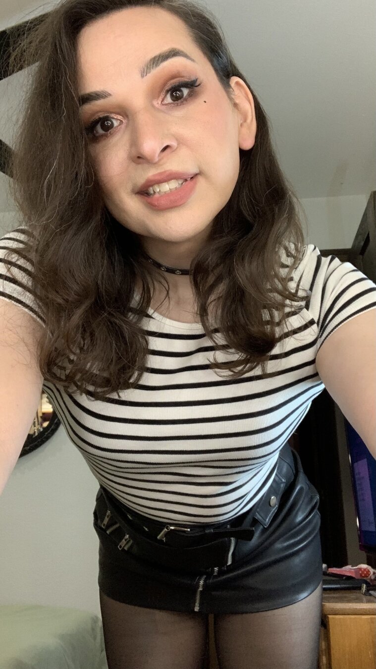 A selfie of a cute trans woman (Bijou) making eye contact. She has wavy long dark brown hair, and is wearing a striped top, a black mini skirt, and black nylons. Room interior.