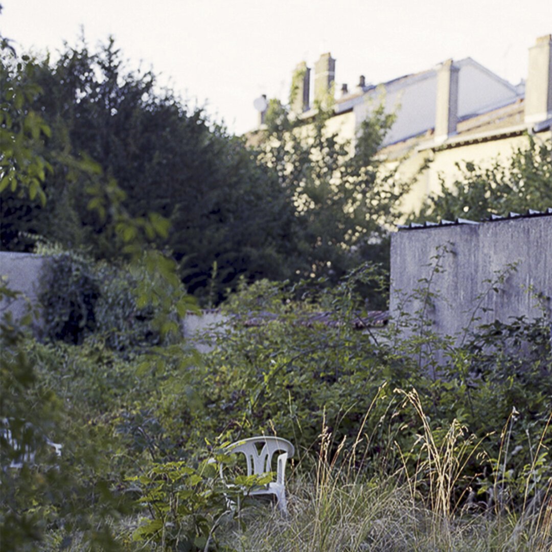 Au premier plan un jardin peu entretenu mais agréable. Au milieu au chaise en plastique, et en arrière plan un mur en béton.